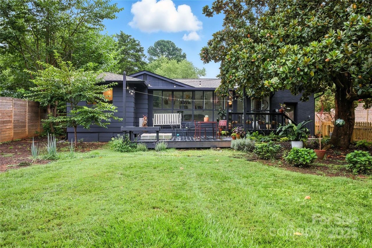 Back view of a dark blue house with large windows and a porch, surrounded by green grass, trees including a large one with broad leaves, and various potted plants under a blue sky.