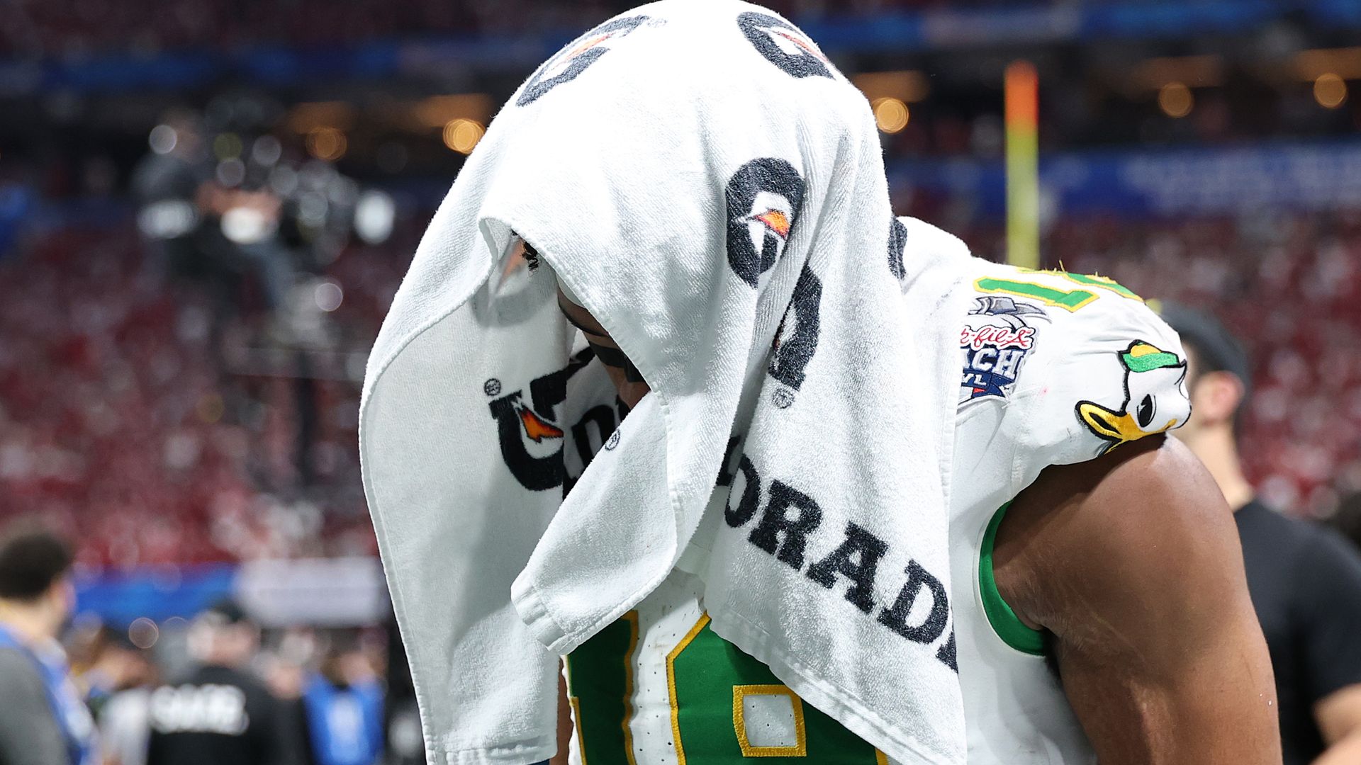 Football player in green and yellow jersey with a white Gatorade towel over their head, appearing dejected on a stadium field with blurred background.