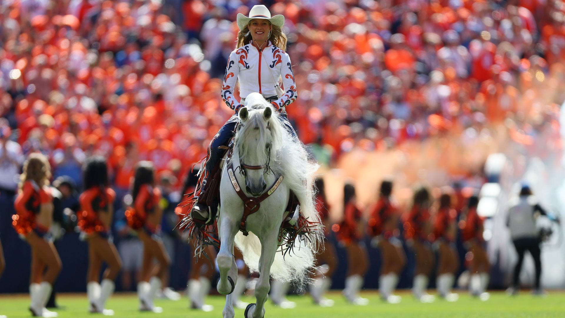 A female rider in a white embroidered jacket and cowboy hat rides a white horse toward the camera, as a sea of orange-clad spectators blur in a crowded stadium behind.