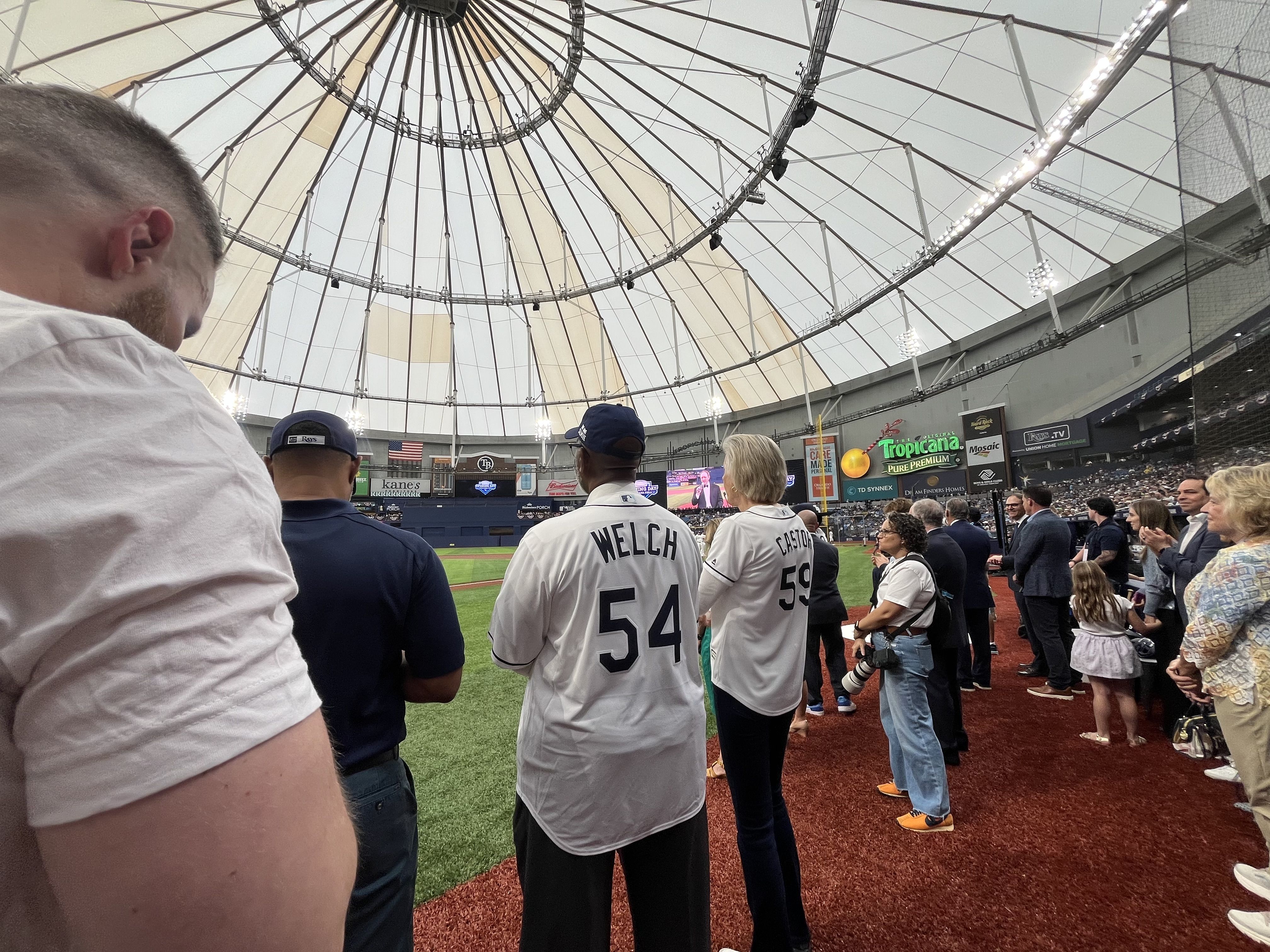 Inside a domed baseball stadium with a circular roof, fans line the red turf along the edge of the field as two players in white jerseys (Welch 54, Castro 59) face the outfield.
