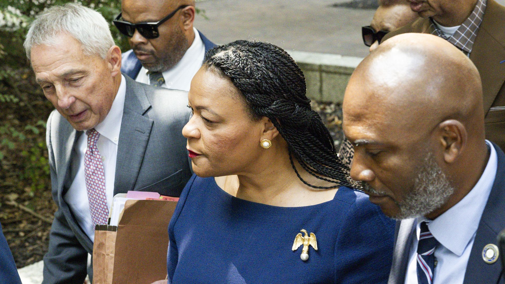 A group of six formally dressed people walking closely together outdoors, including a woman in a blue dress with braided hair and a gold eagle brooch, and men in suits and ties.