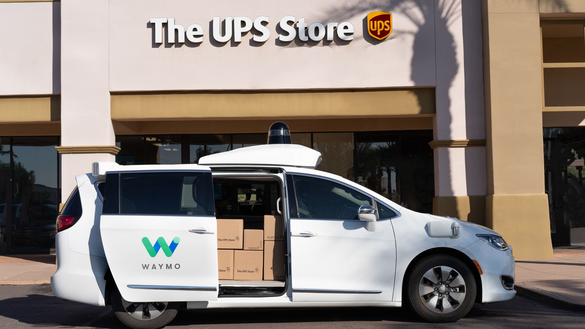 Image of Waymo self-driving minivan loaded with packages in front of a UPS store.