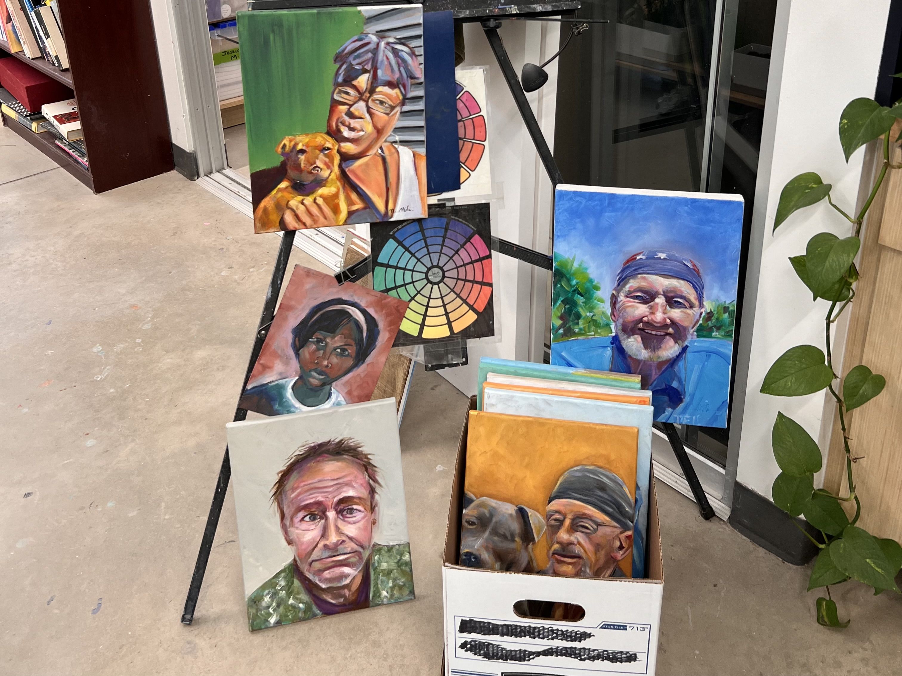 Colorful portraits displayed on a metal stand in a shop: a woman with glasses holding a yellow dog, a color-wheel artwork, a man with a blue bandana, and other vibrant faces; a plant sits to the right.