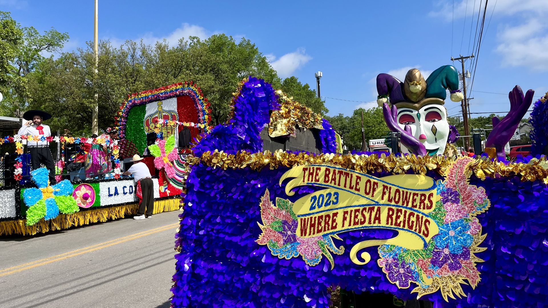 A purple, Mardi Gras like float that says "The Battle of Flowers 2023, Where Fiesta begins."