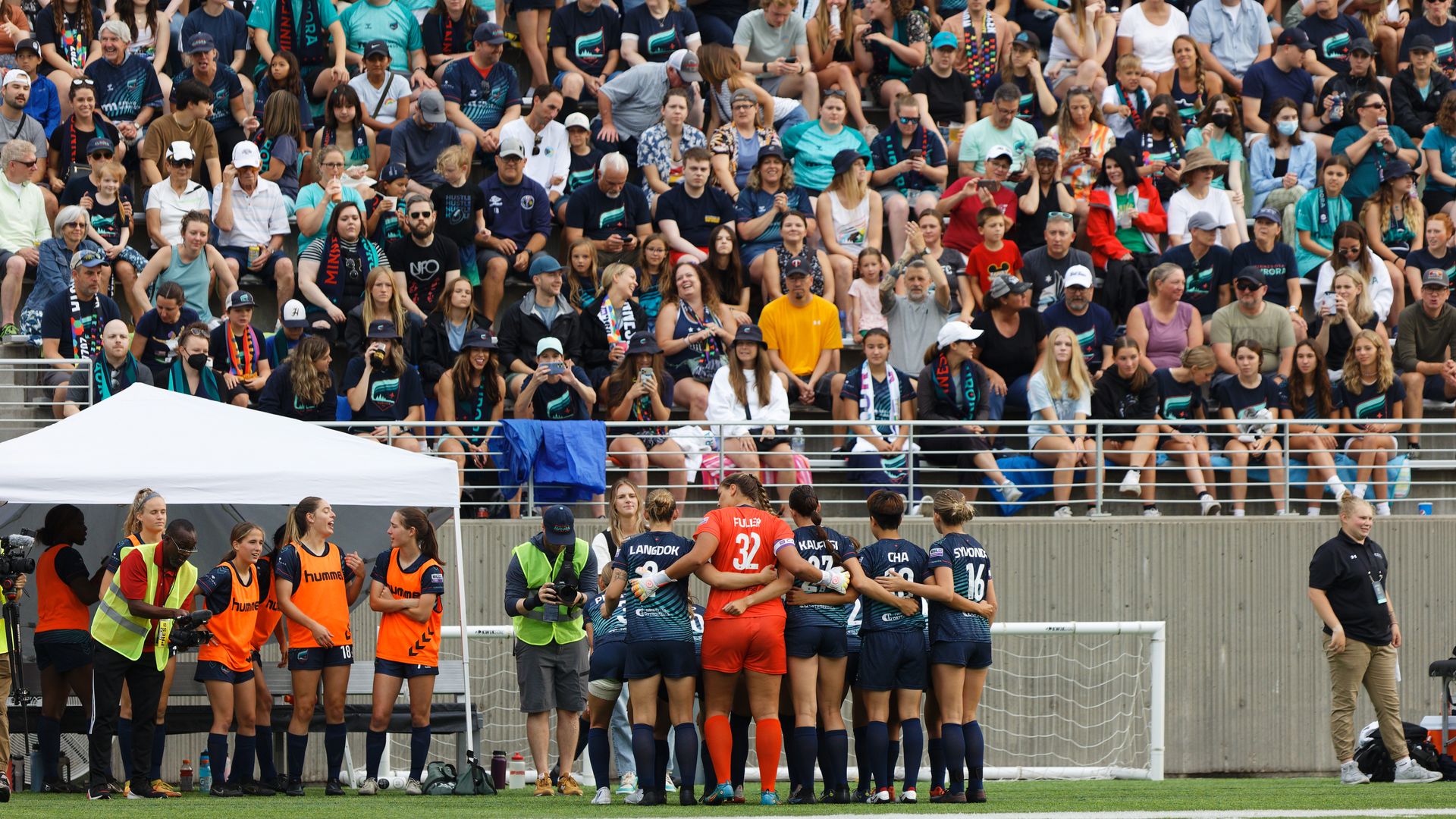 A packed stadium with fans in teal scarves watching a match; foreground shows players in blue and navy near a goal on green turf, while photographers and staff in orange vests observe.