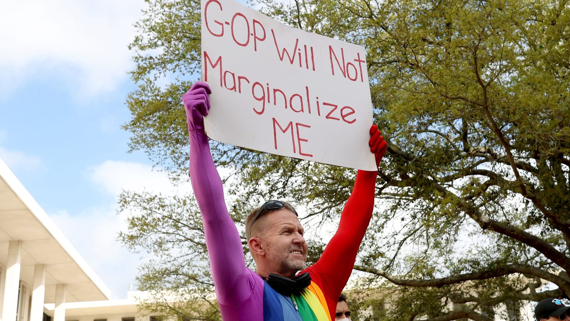A person in a rainbow bodysuit holds up a sign reading "GOP Will Not Marginalize ME"