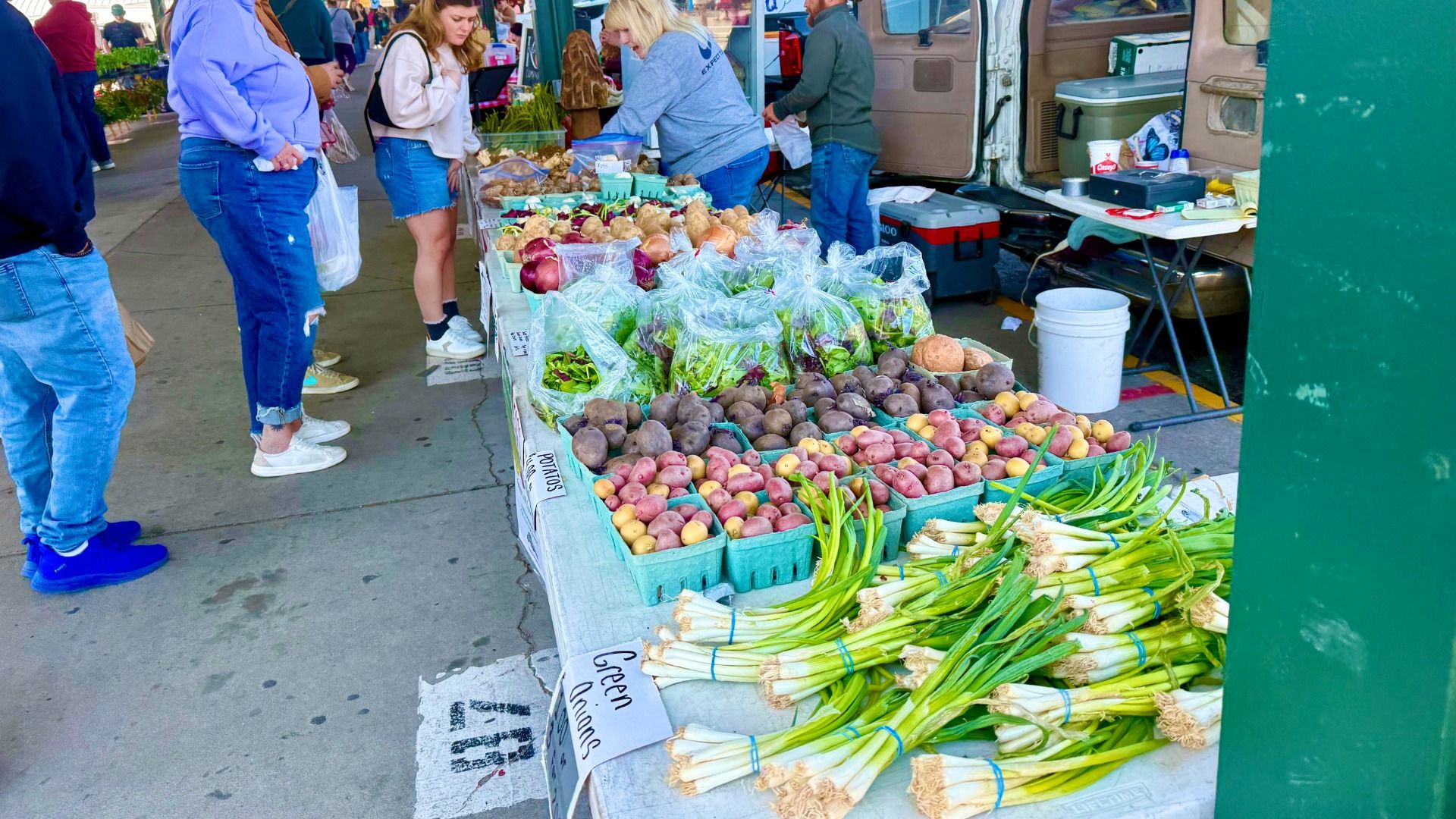 Outdoor market scene with a long vegetable stall: leeks and greens in the foreground, potatoes and onions on display; shoppers in jeans and hoodies browse as a van with open doors sits at the back.