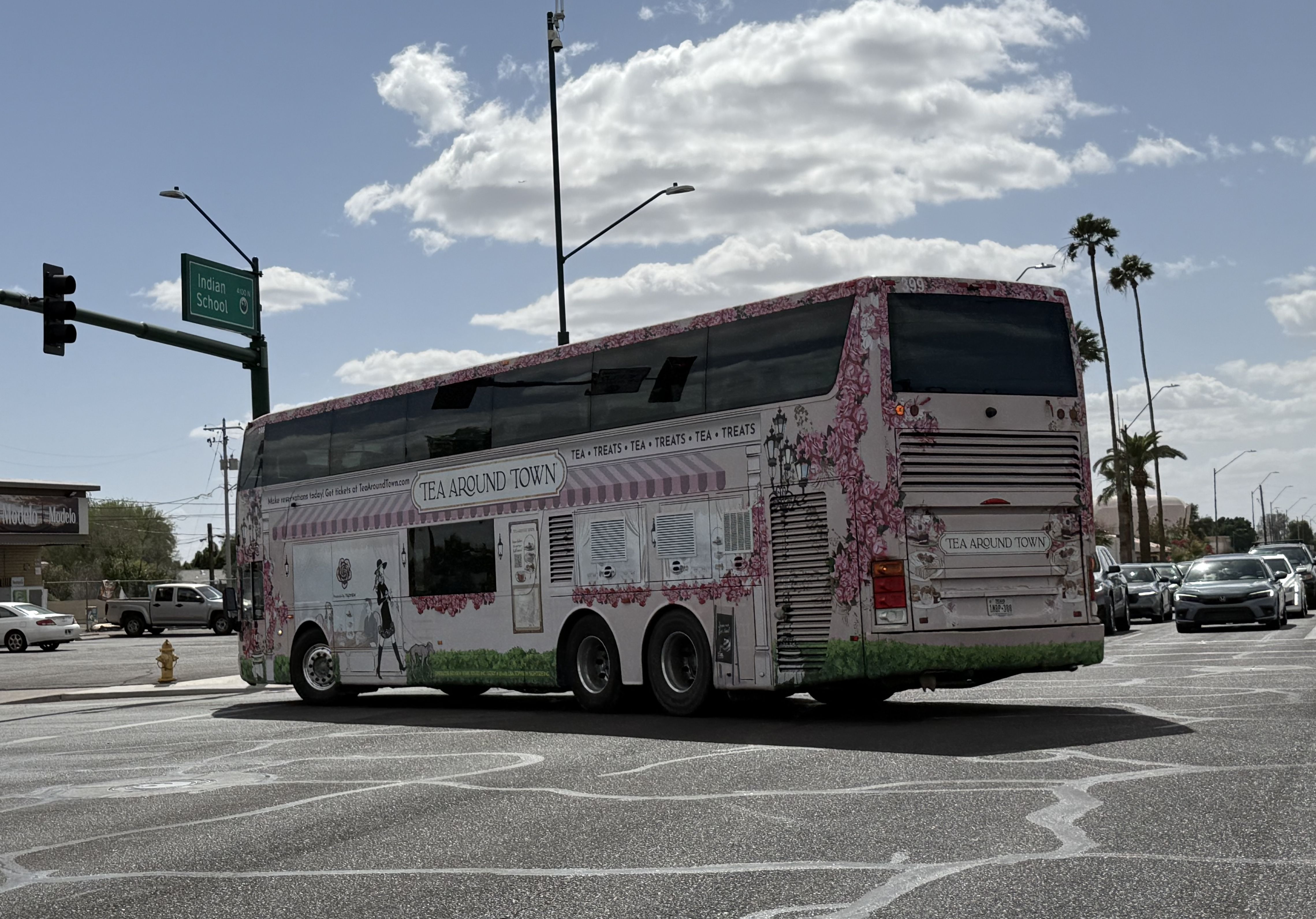 Pink floral tour bus with "Tea Around Town" signage turns left through an intersection, near a green street sign that reads Indian School and tall palm trees.