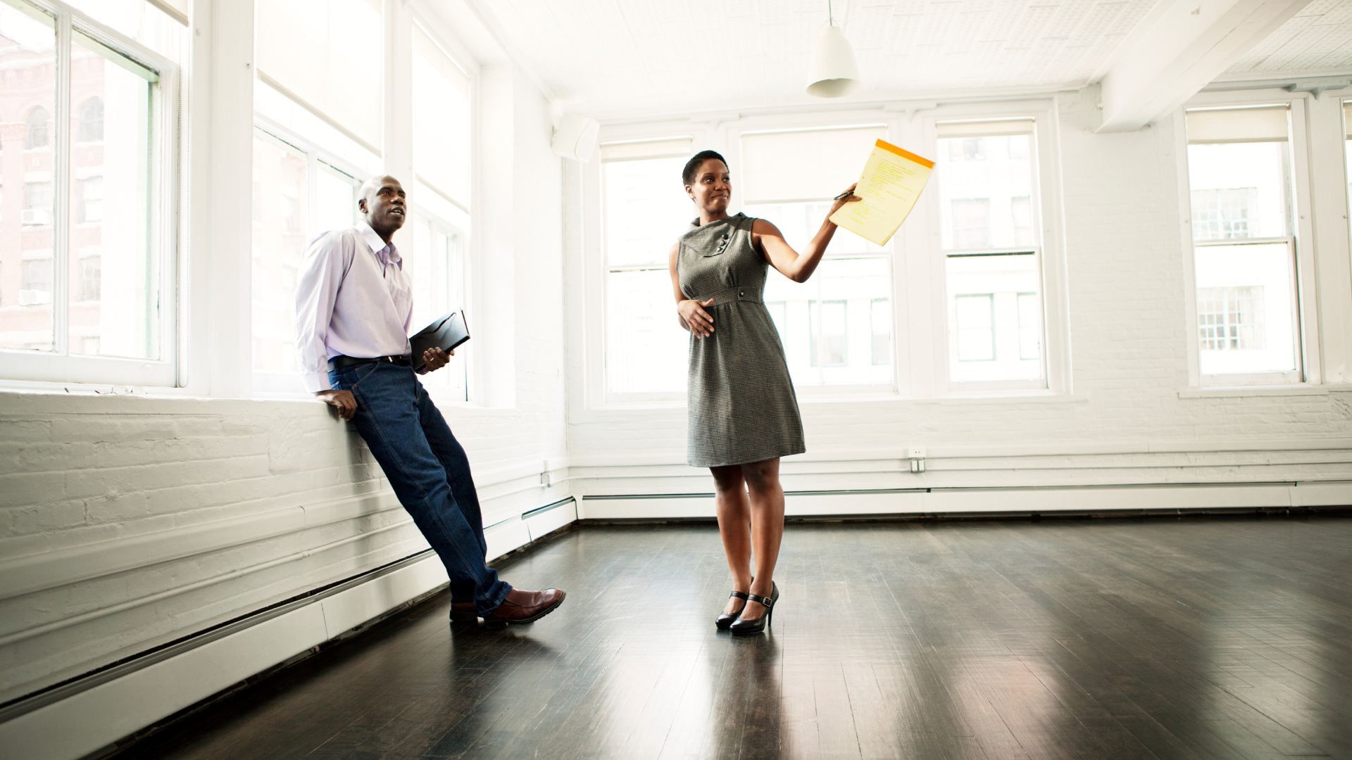 Two professionals in a bright room: a man in a lavender shirt and jeans leans on a windowsill with a notebook, while a woman in a gray dress holds a yellow clipboard.
