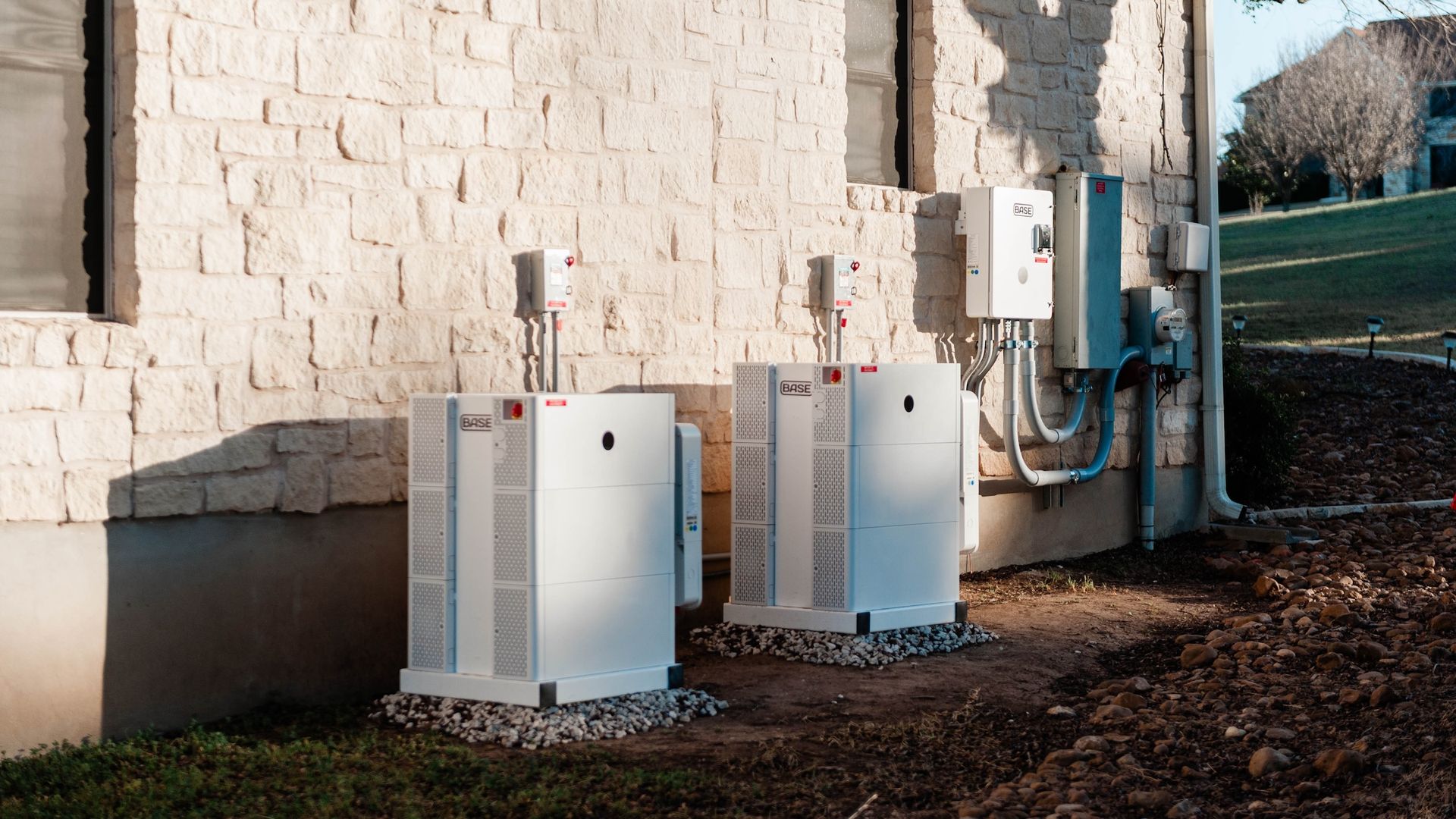 Two white BASE battery storage units installed outside a beige stone building wall with electrical panels and meters, set on gravel with grass and rocks nearby under evening sunlight.