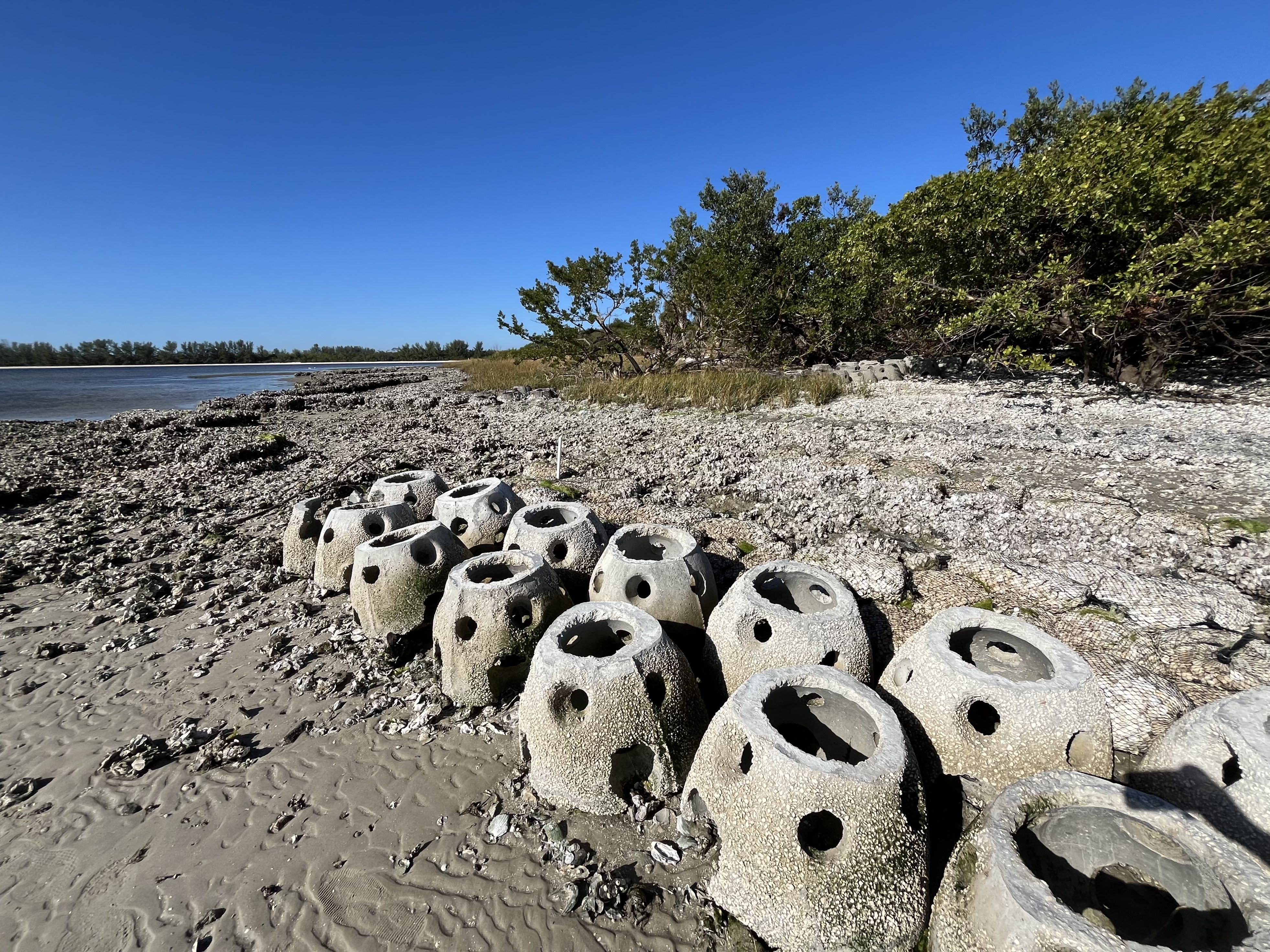Domes of concrete with holes in them resting on a sandy shoreline with trees and brush in the background.