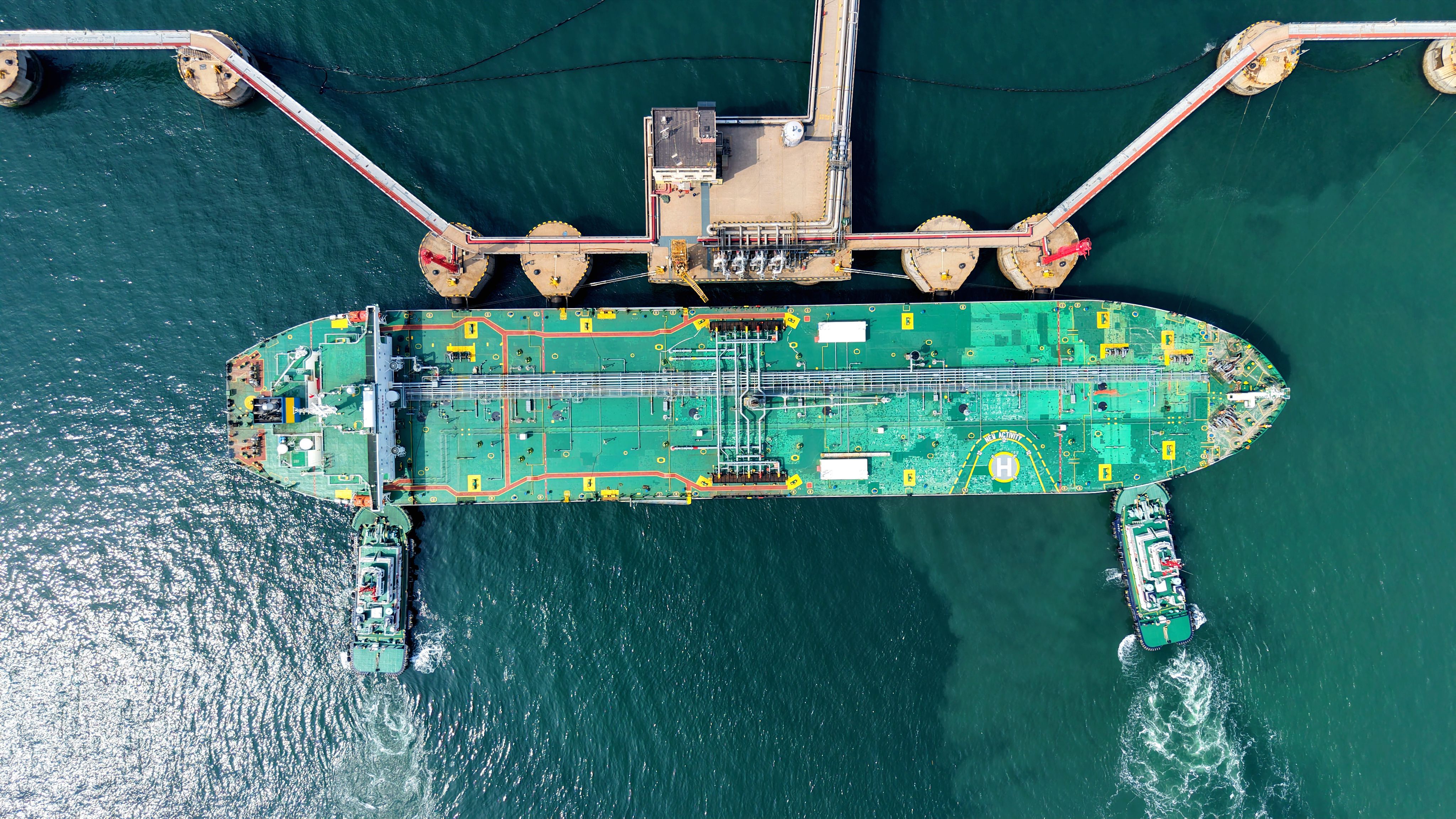 A large oil tanker docked at an industrial terminal in Qingdao Port, with pipelines and infrastructure used for unloading crude oil visible along the waterfront.