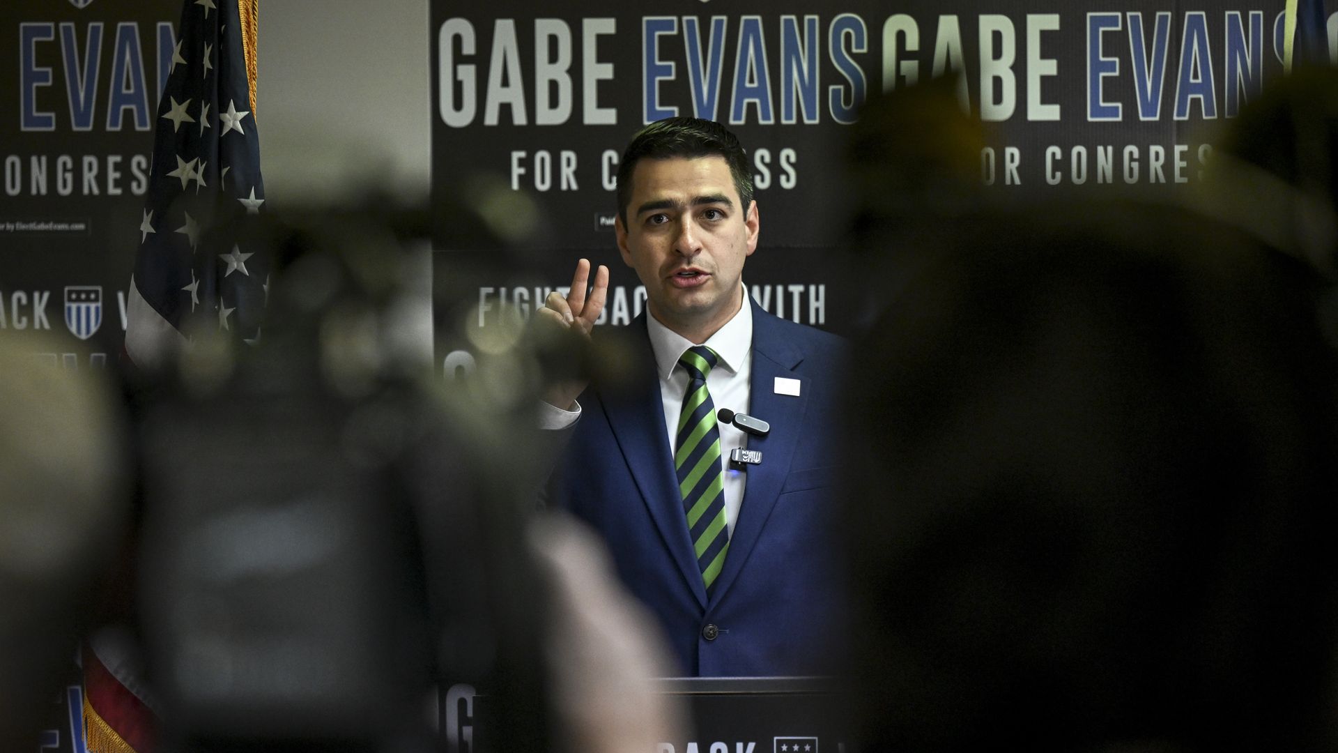 Man in a blue suit and green-striped tie speaks at a podium with American flag and campaign signs reading "Gabe Evans for Congress" in the background.