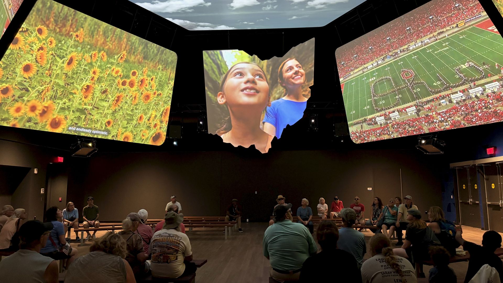 Inside a dark room, three large angled screens show left a sunflower field, center two smiling girls, and right a packed OSU football stadium with Script Ohio; audience sits on benches.