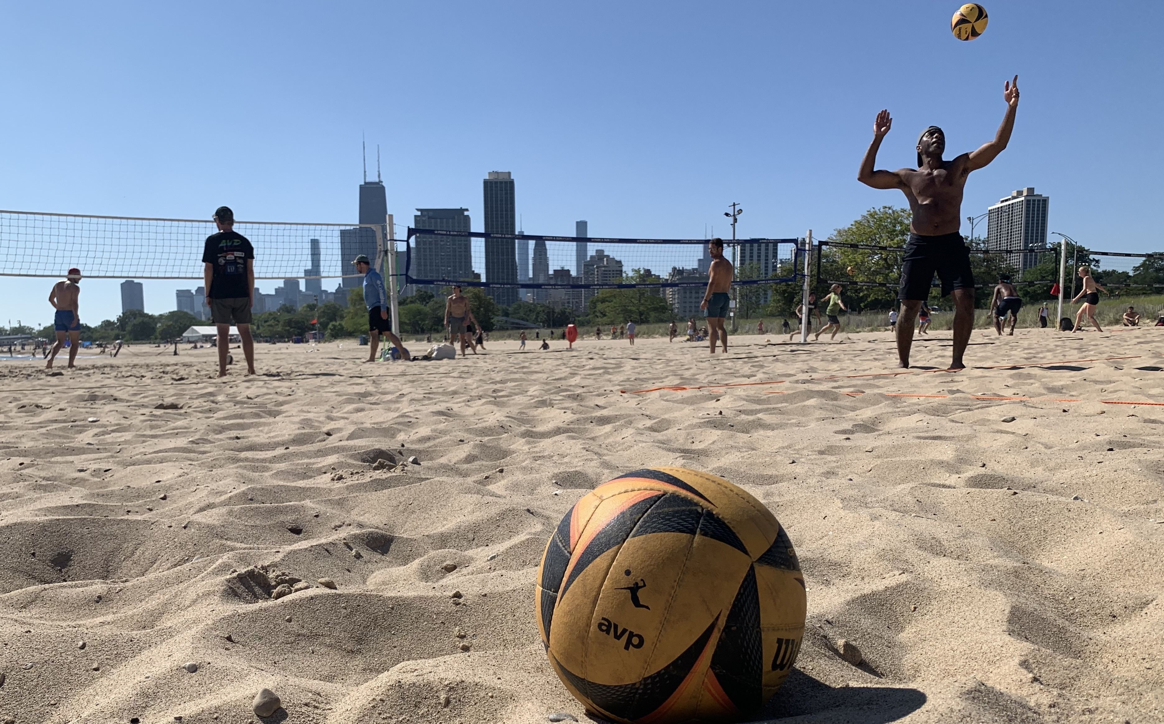 Sunlit beach volleyball game on a sandy court; players near a net, one shirtless man jumping; a yellow and black AVP volleyball sits in the foreground, with a city skyline against a clear blue sky.
