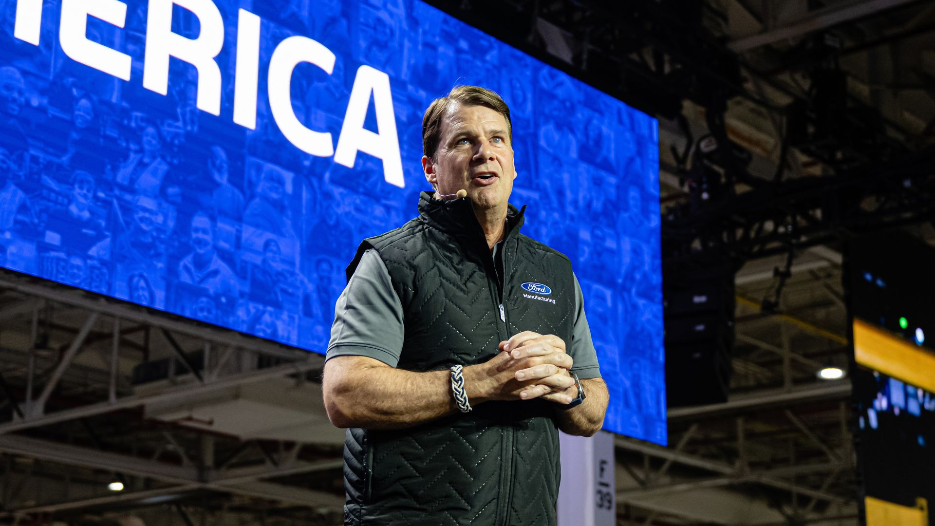 Ford CEO Jim Farley, wearing short sleeves and a black manufacturing vest, speaking on stage at a Ford factory in Louisville, KY