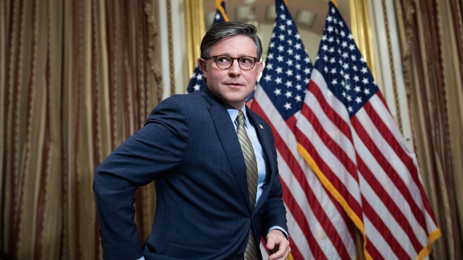 A man in a blue suit and glasses stands in front of several American flags in a formal room with ornate gold-trimmed curtains.