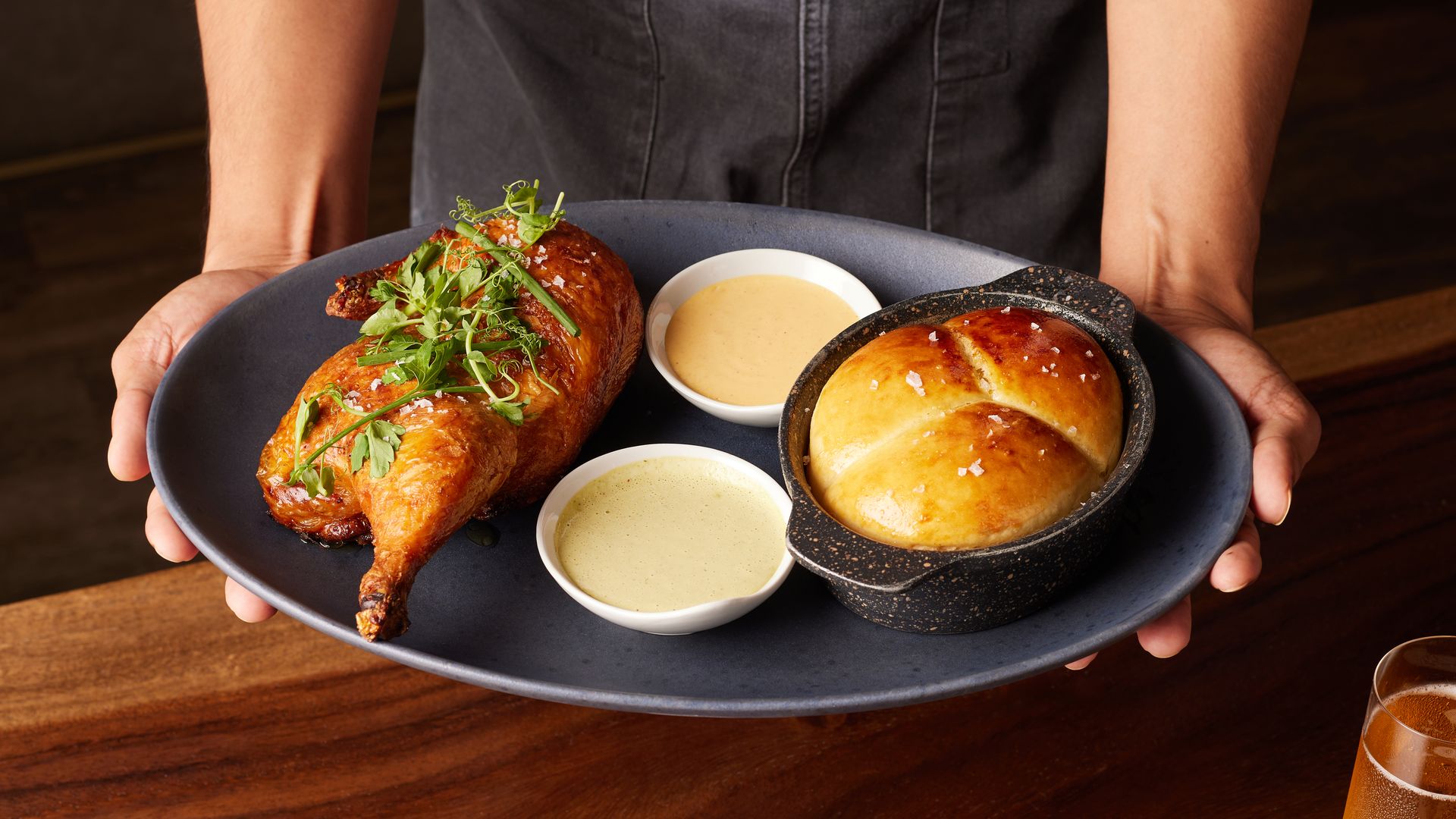 Two hands hold a black platter with a roast chicken, milk bread roll and sauces 