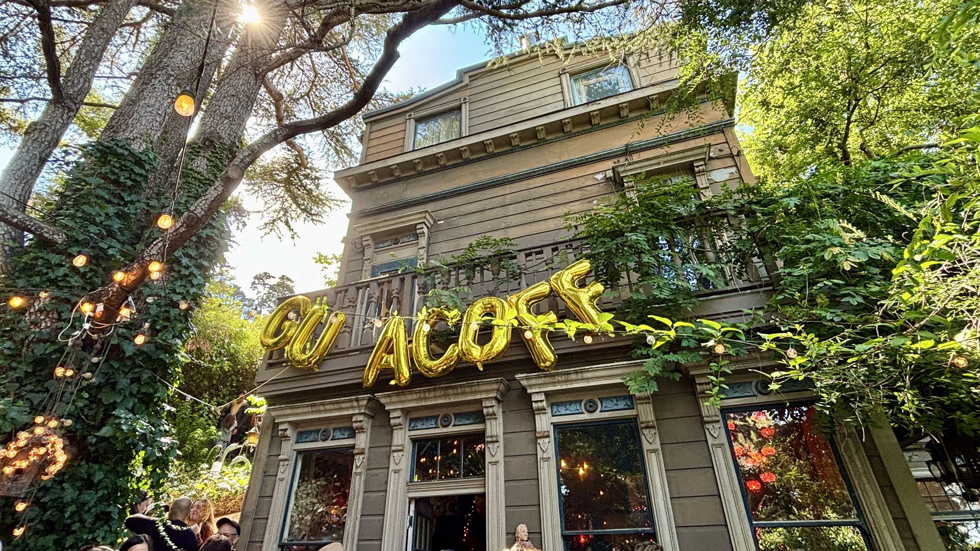 Outdoor party at a rustic house with greenery and string lights. Gold balloons spell out part of the word "GUACOFF" above the entrance under a sunny sky.