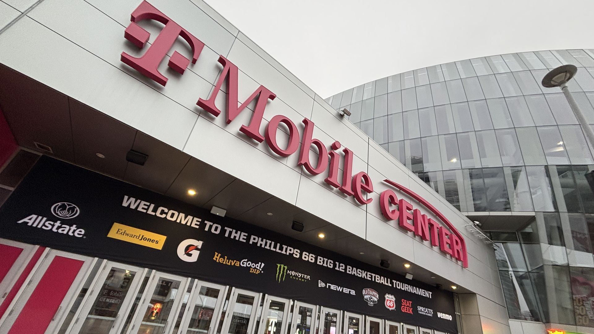 Exterior of T-Mobile Center with large magenta signage, glass building in background, and a black banner welcoming visitors to the Phillips 66 Big 12 Basketball Tournament with sponsor logos.