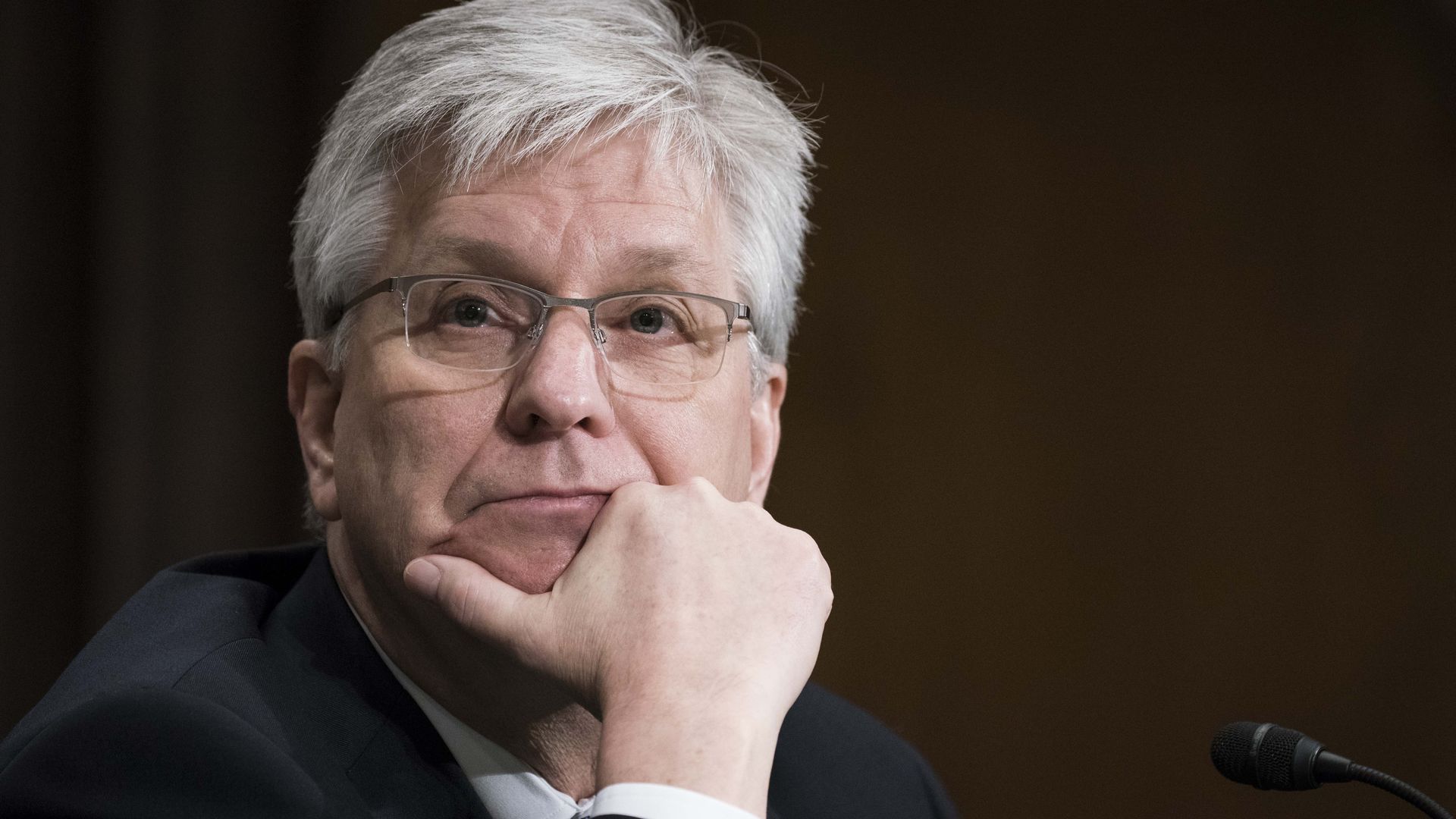 Fed governor Christopher Waller, with white hair and glasses, wearing a dark suit, resting his chin on his hand, sitting near a microphone against a dark background.