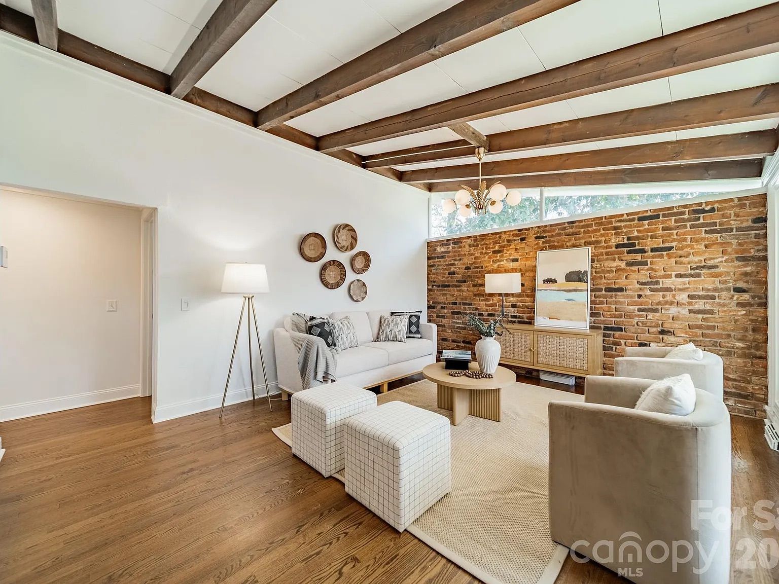 Cozy living room with white walls, exposed dark wood beams, and a brick accent wall. White sofa, two checkered ottomans, round coffee table, beige rug, and art on the brick wall.