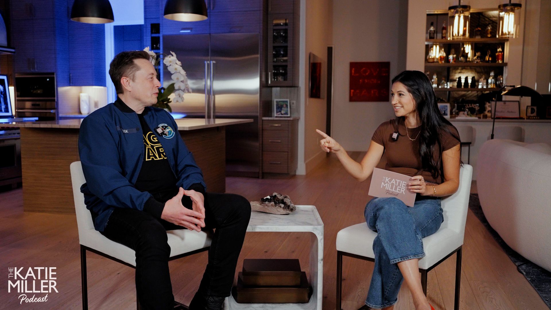 Elon Musk, wearing a blue jacket and black pants talks with Katie Miller, wearing a brown top and jeans holding a card that says "The Katie Miller Podcast" in a modern, warmly lit room.