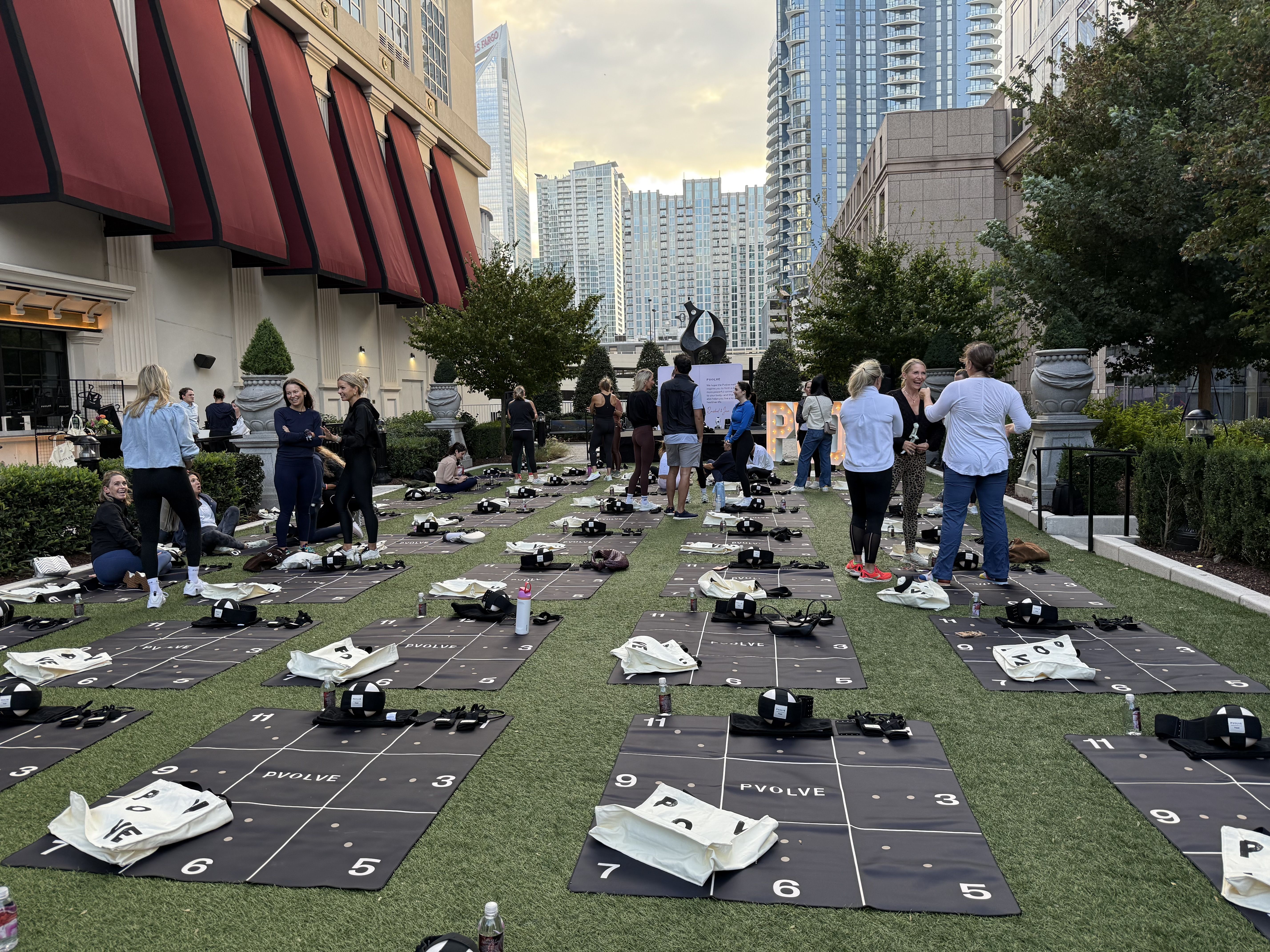 Outdoor fitness event with black PVOLVE mats and gear arranged on green grass, people in activewear chatting, tall buildings and trees surround the area under a cloudy sky.