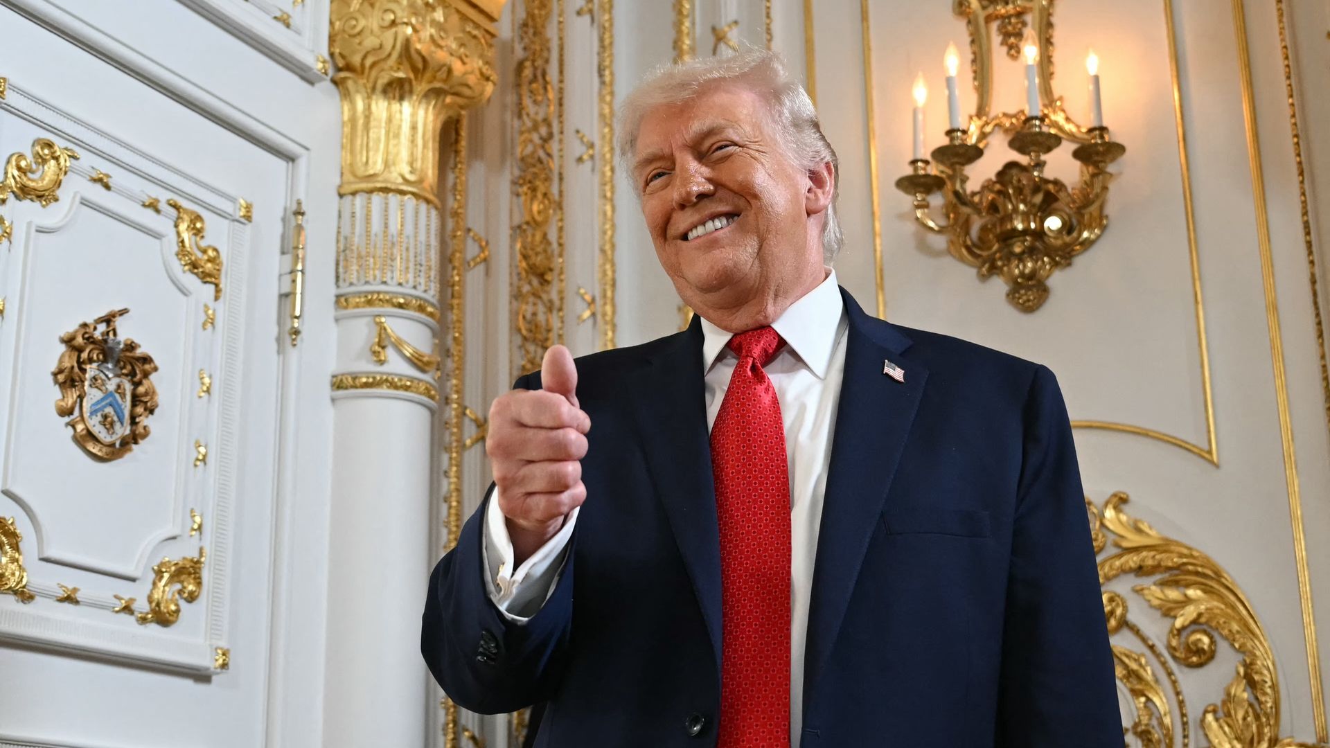 Smiling Donald Trump in a navy suit, white shirt, and red tie giving a thumbs-up in an ornate room with white and gold decor and a lit wall sconce.