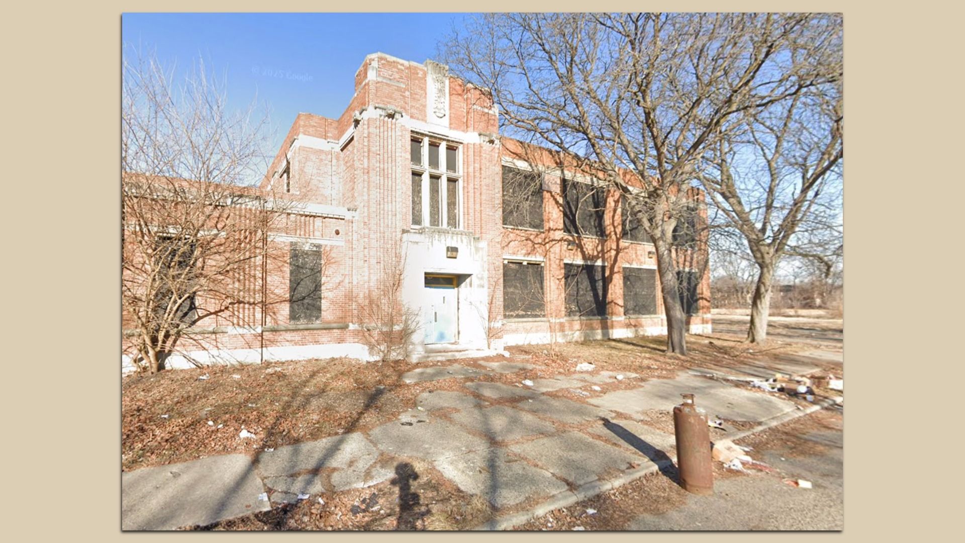 Abandoned red-brick two-story building with tall windows, peeling stone trim, surrounded by leafless trees. Cracked pavement and scattered debris.