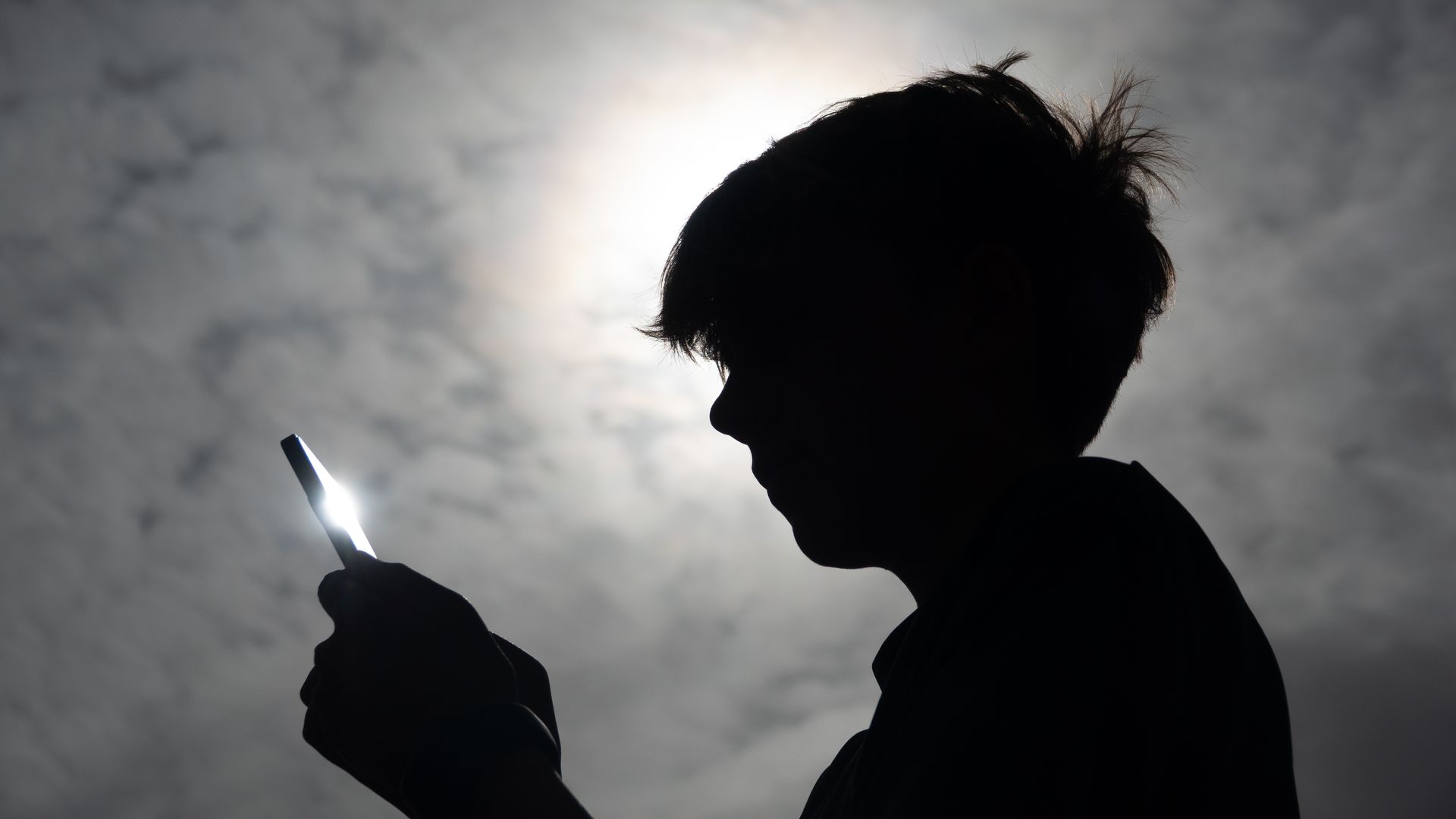 The silhouette of a 13-year-old teenage boys looking at his iPhone smart phone screens on August 26, 2025 in Penzance, England, beneath a cloudy sky as the sunshine breaks through/