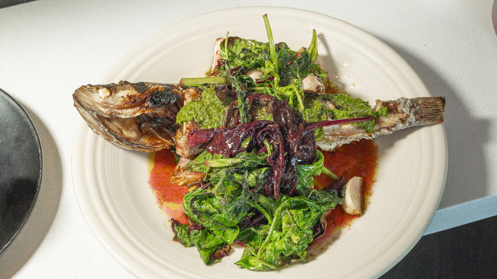 Whole grilled fish placed across a bed of sautéed greens (purple and green leaves) on a white plate, with red sauce pooled beneath.