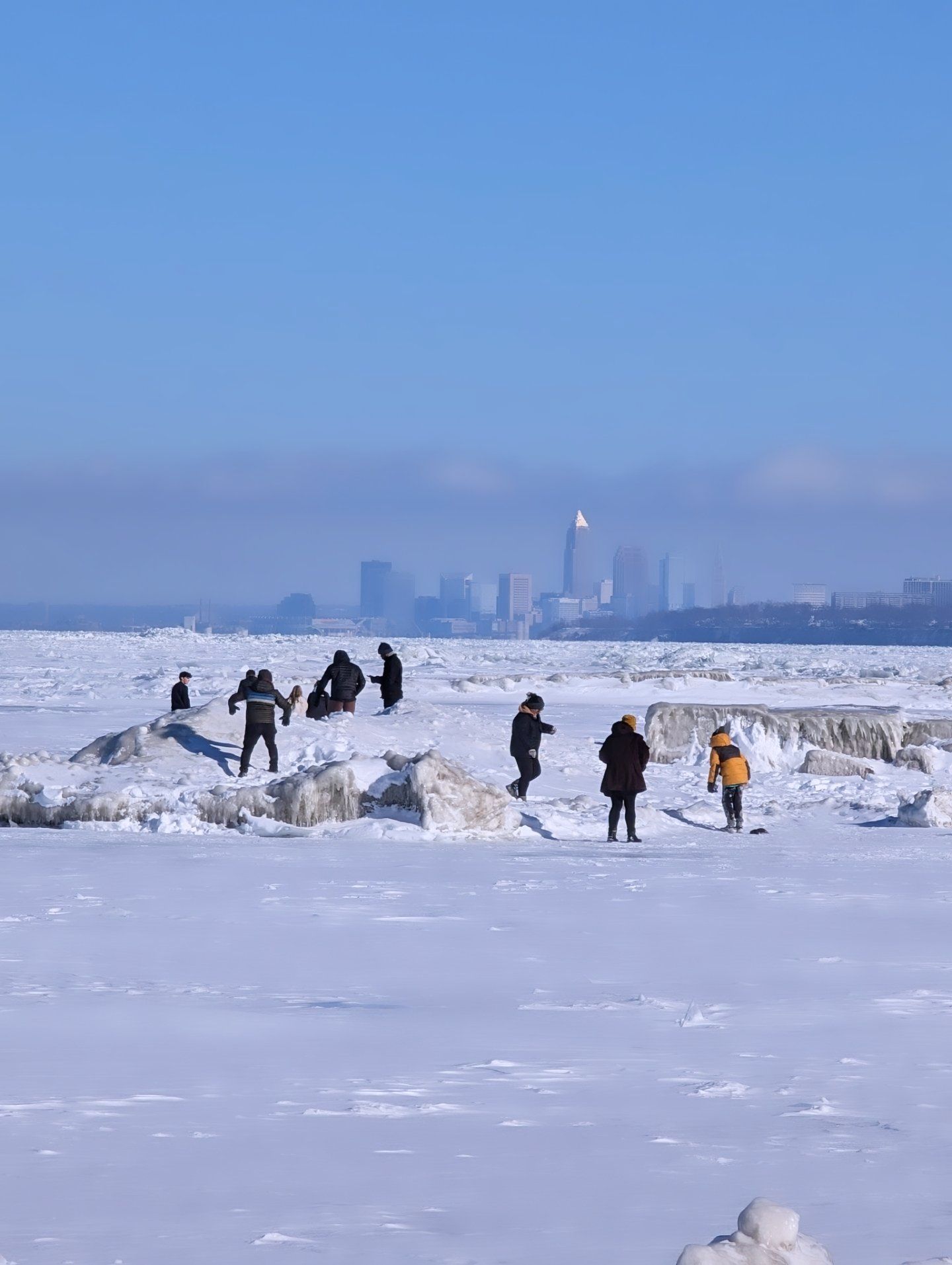 People dressed in winter clothing walk and play on a snowy frozen lake with large ice chunks under a clear blue sky, city skyline visible in the background.