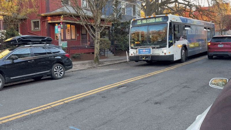City bus labeled “15 NW 23rd Ave” stopped on a residential street with parked cars. A red house with a porch and bare trees line the sidewalk, suggesting autumn or winter in a quiet neighborhood setting.