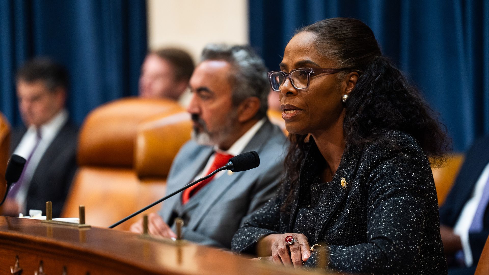Del. Stacy Plaskett, wearing a black and white blazer, speaks into a microphone at a wooden dais.