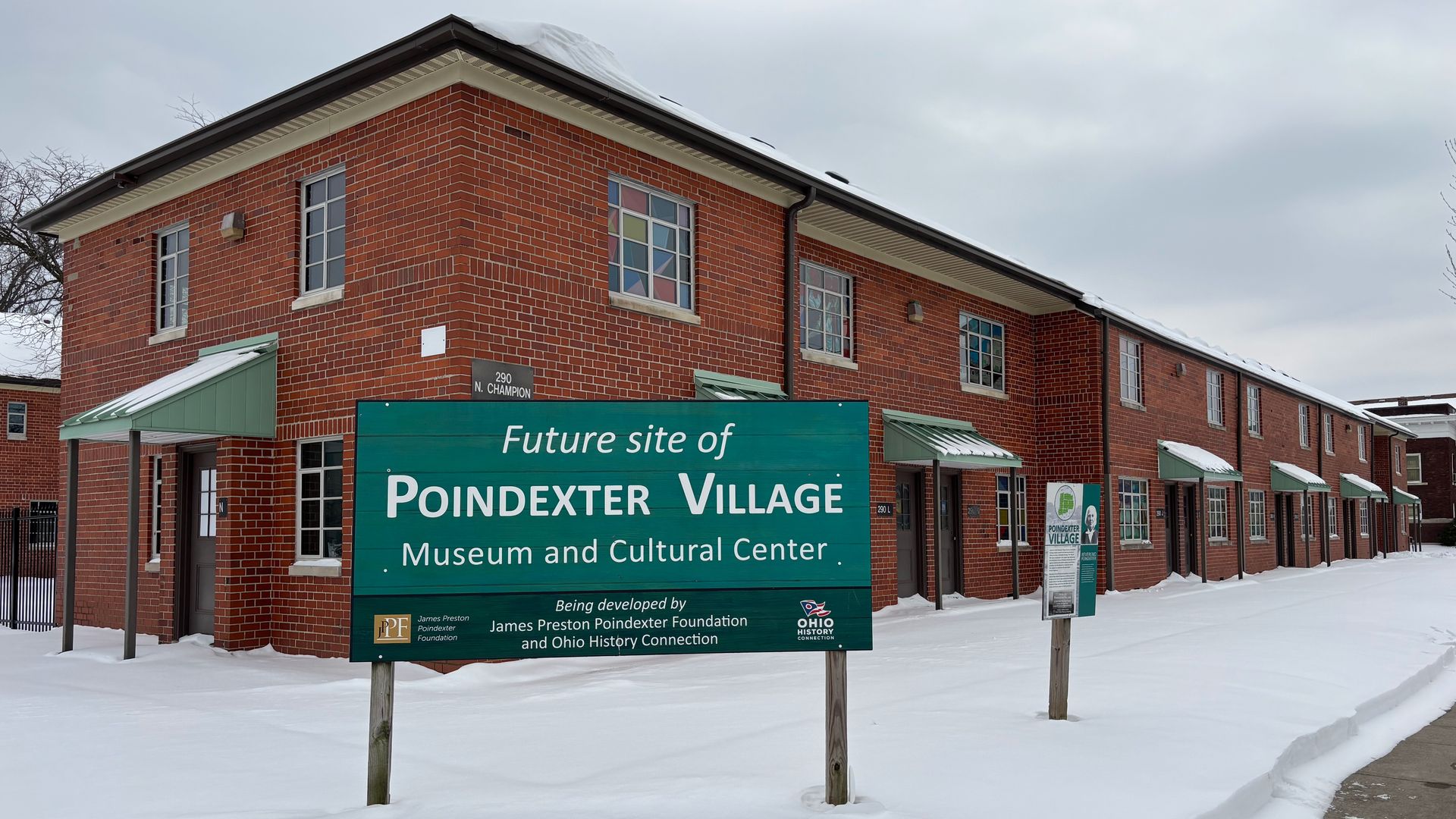 Snow-covered brick building with green awnings and signs announcing the future site of Poindexter Village Museum and Cultural Center in cloudy weather.