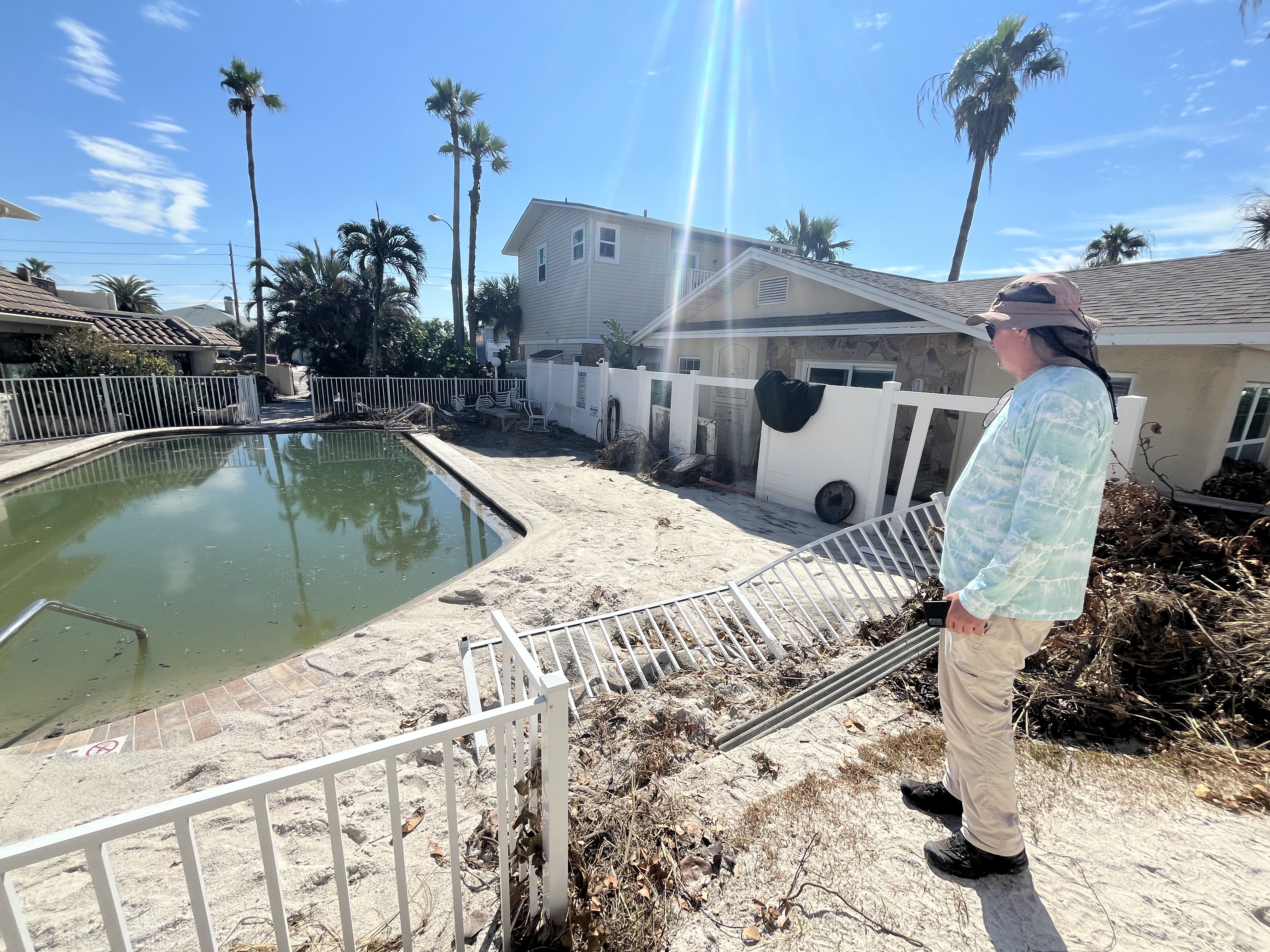 A man in khaki pants, a blue rashguard and a safari hat looks out over a greenish-brown swimming pool surrounded by feet of sand.