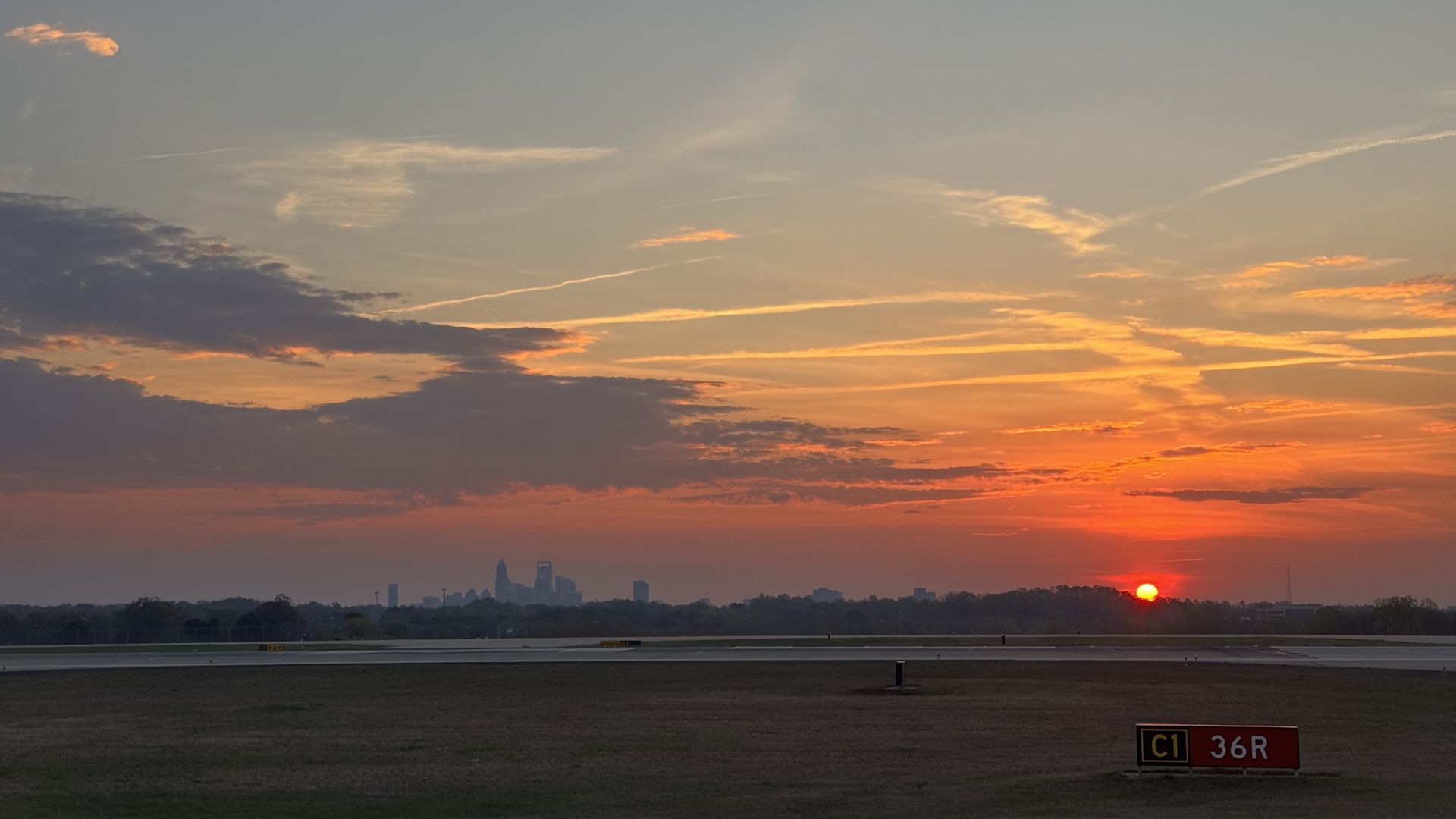 Sunset over an airport runway with orange and pink clouds, a distant city skyline on the horizon, and a red sun near the lower right; foreground shows grass and a C1 36R sign.