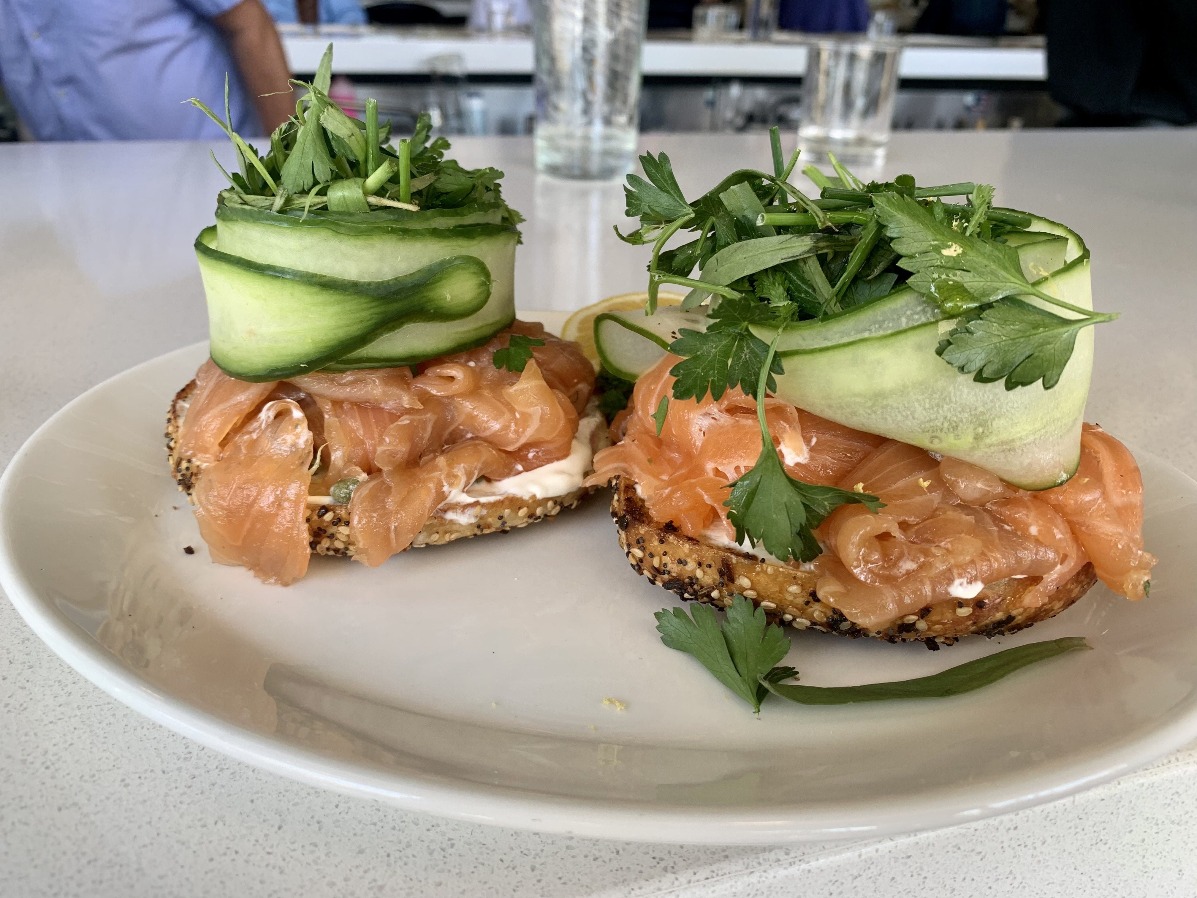 a giant stack of smoked salmon on a bagel topped with cucumber