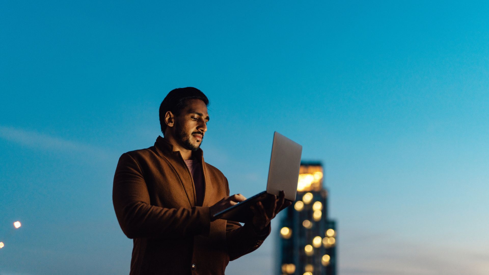 Man in a brown jacket stands outdoors at dusk, looking down at an open laptop he holds. A blue evening sky and blurred city lights glow in the background.