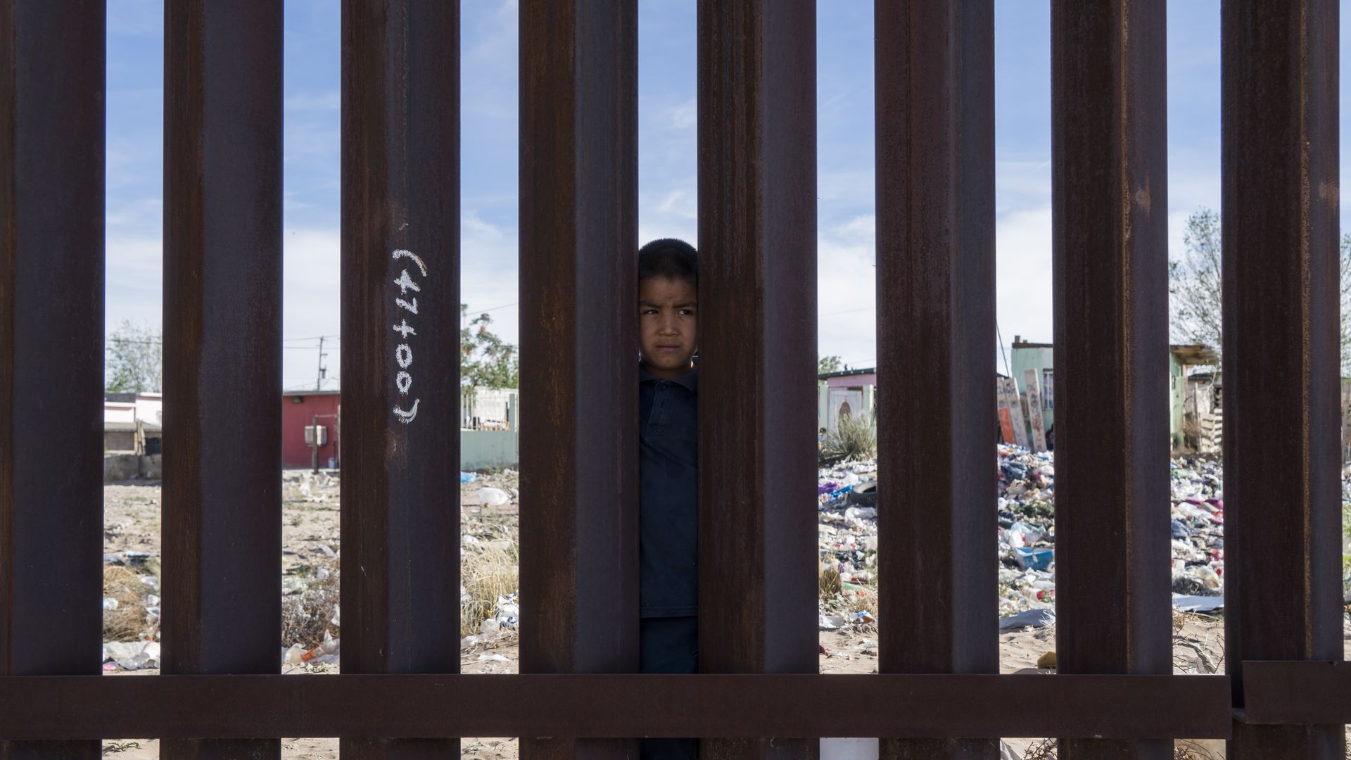 Little boy stand on the Mexico side of the border wall with his head peeking through the slats of the metal fence.