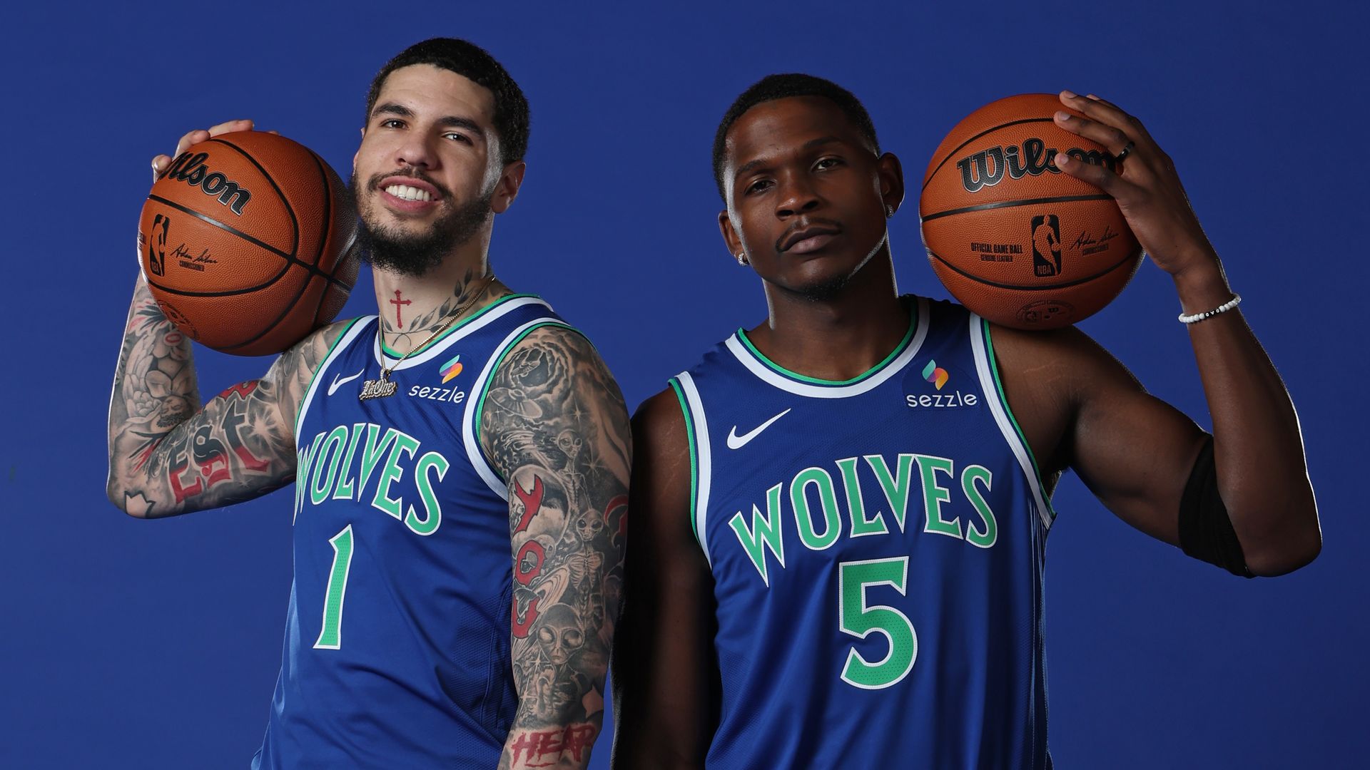 Two Wolves basketball players stand back-to-back in blue jerseys against a blue backdrop, each holding a basketball on their shoulders. Player 1 has tattoos; Player 5 wears light-blue sneakers.
