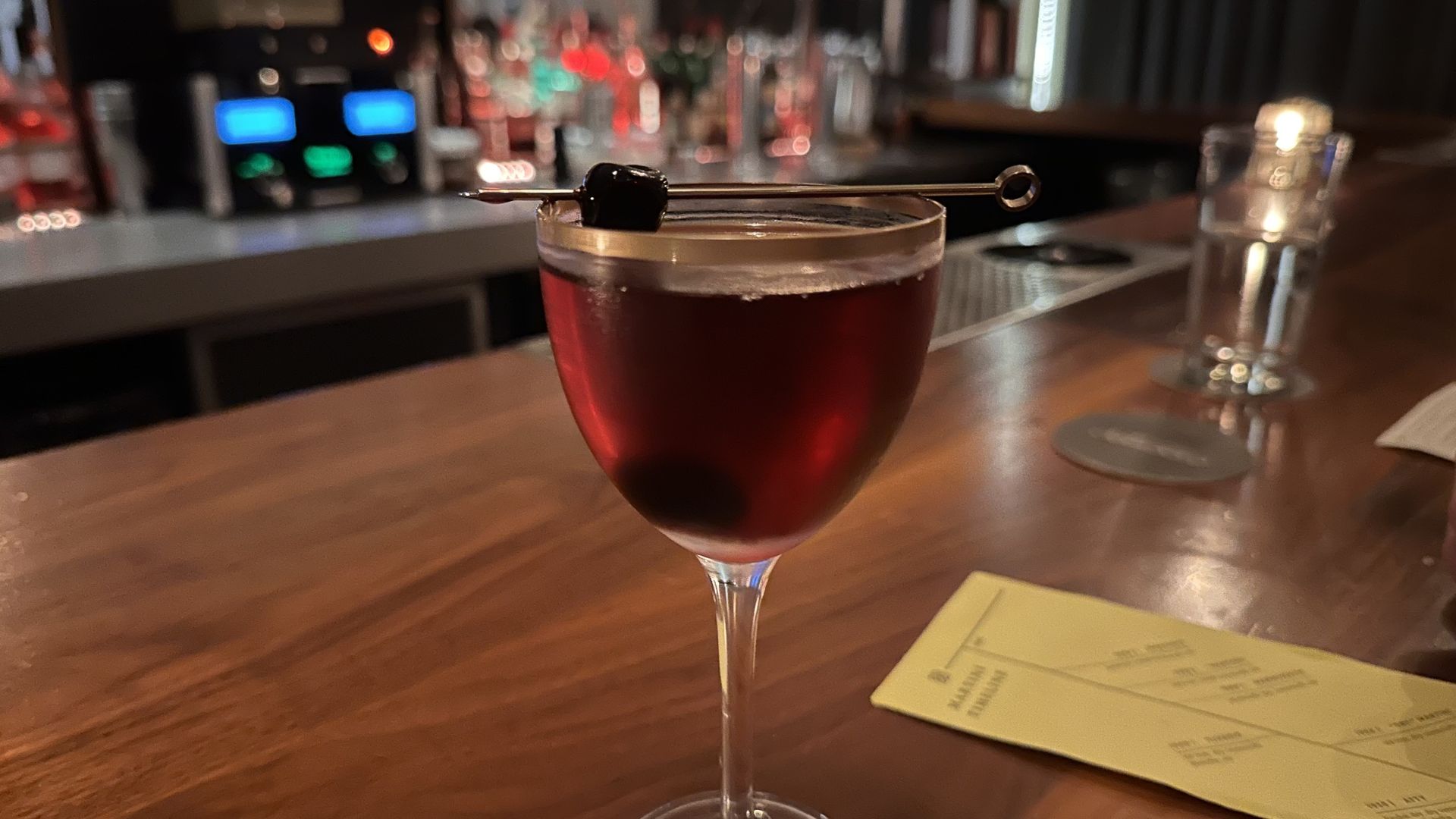 A dark red cocktail in a clear glass on a wooden bar counter, garnished with a black cherry on a skewer, with a yellow menu and a glass of water in the background.