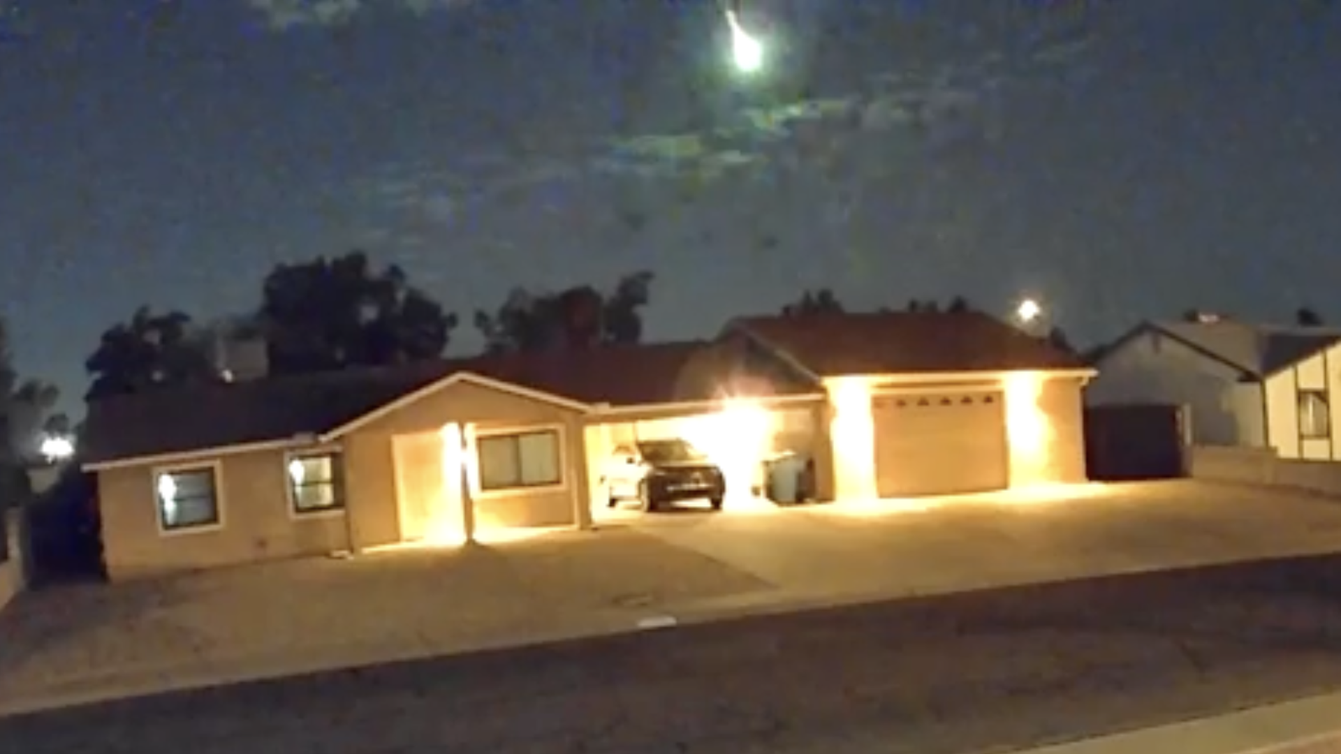 Nighttime view of a beige, single-story house with a two-car garage. A car sits in the open garage; warm exterior lights illuminate the driveway, with trees and neighboring homes in the background. A green meteor explodes in the sky above.