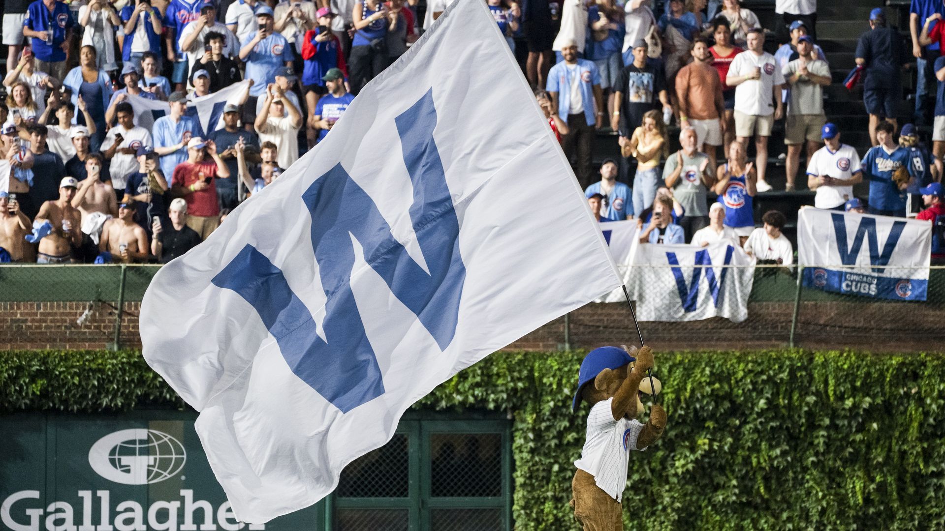 Wide crowd of baseball fans in blue and white fills the stands as a large white flag with blue geometric shapes waves in front; a mascot and Gallagher Insurance banners are visible.