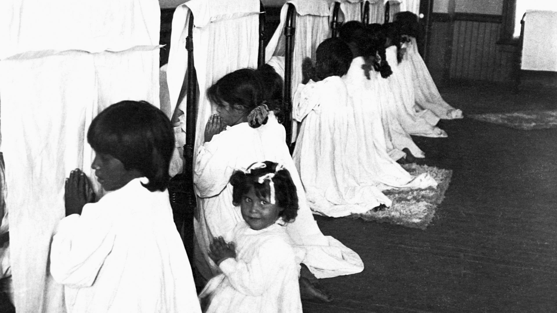 A balck-and-white image of children praying and one girl looking at the camera. 