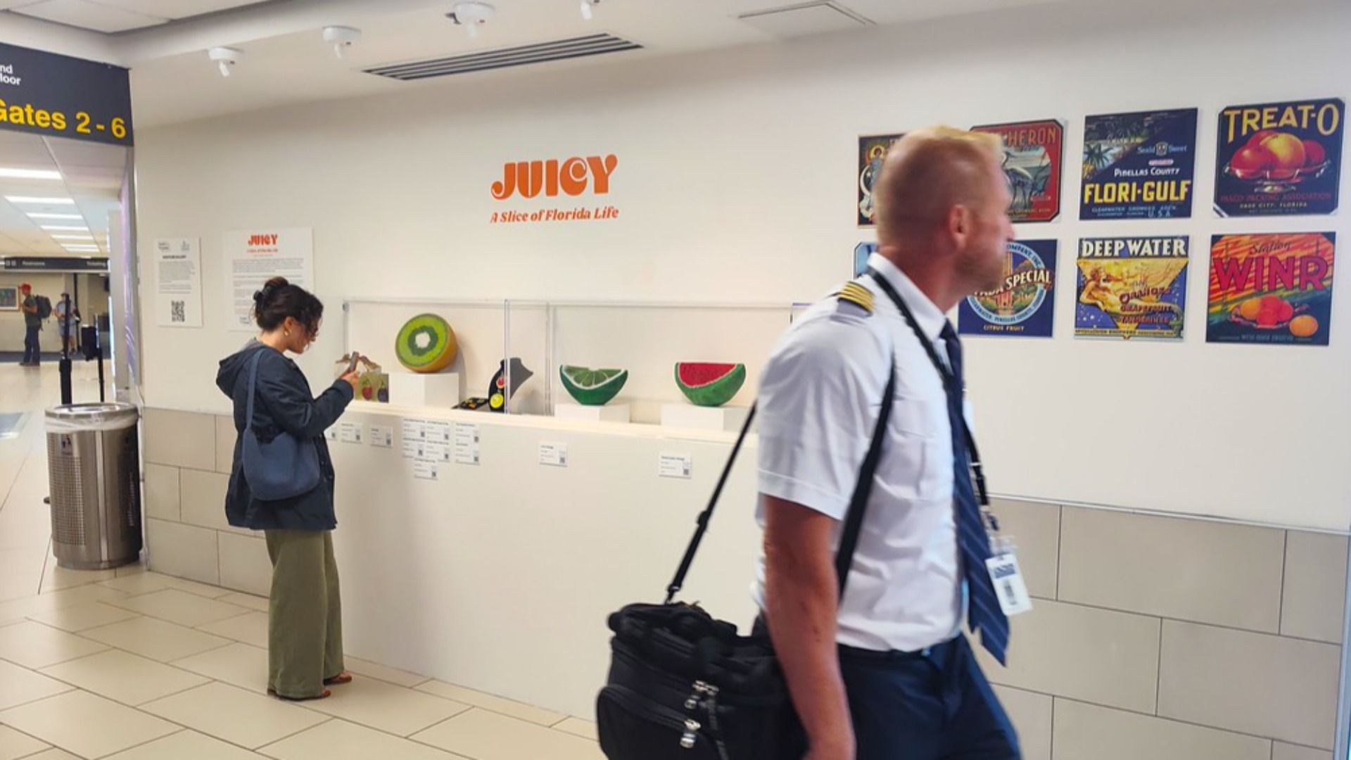 Airport interior with a white wall and orange JUICY logo; a uniformed pilot walks by a fruit display as a woman browses. Colorful travel posters line the right wall; Gates 2-6 sign left.