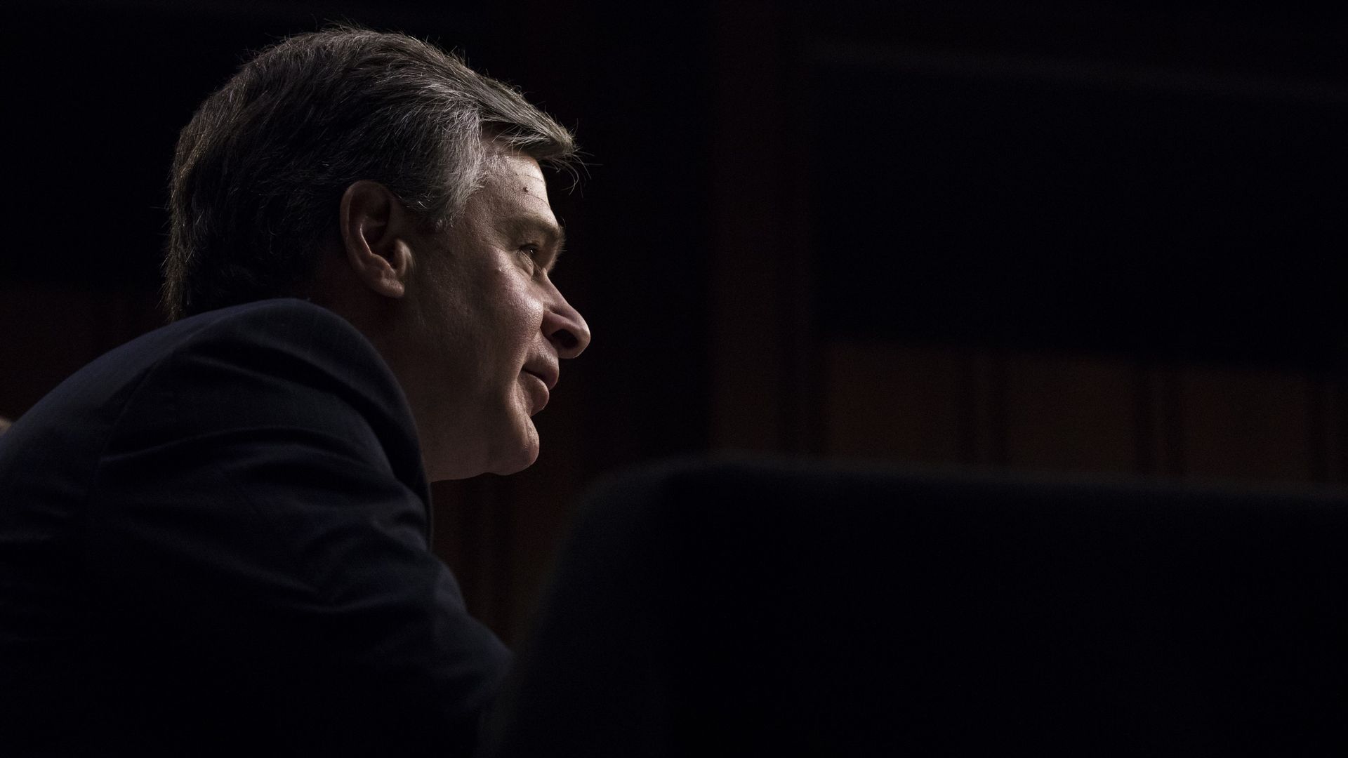 FBI Director Chris Wray in nearly all black, lights above illuminate his face.