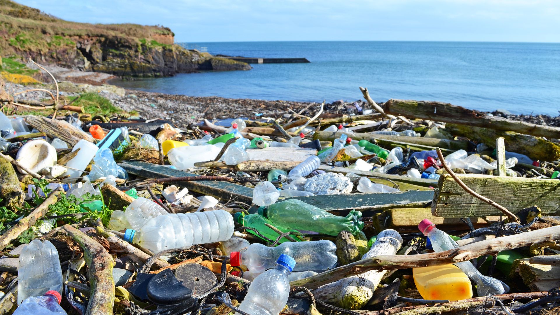 In this image, piles of bottles and other trash are seen on a beach near a cliff.