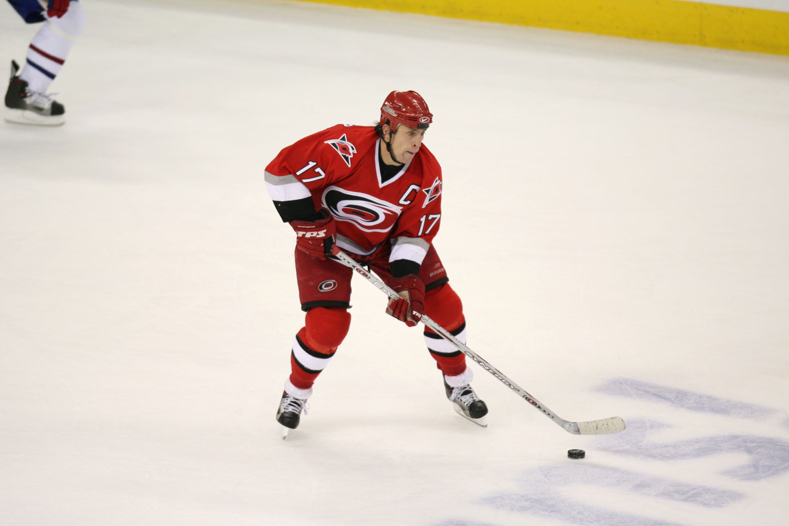 A hockey player wearing a red Carolina Hurricanes uniform with the number 17 and captain’s “C” on the jersey is skating on the ice while handling the puck with his stick during a game.